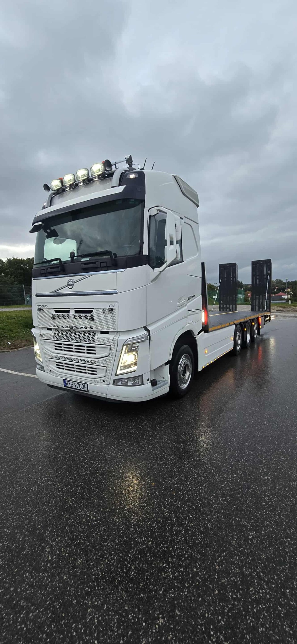 Biała ciężarówka Volvo FH z niskopodwoziową naczepą, oświetlona dodatkowymi lampami, stoi na mokrym asfalcie w pochmurny dzień. Widok z przodu, lekki kąt.