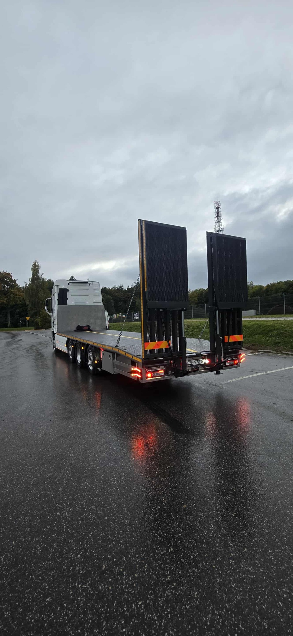 Nisko zawieszona laweta z podnoszonym najazdem, widziana z tyłu na mokrym asfalcie. Widoczne światła stopu i tablica rejestracyjna. W tle drzewa i pochmurne niebo.