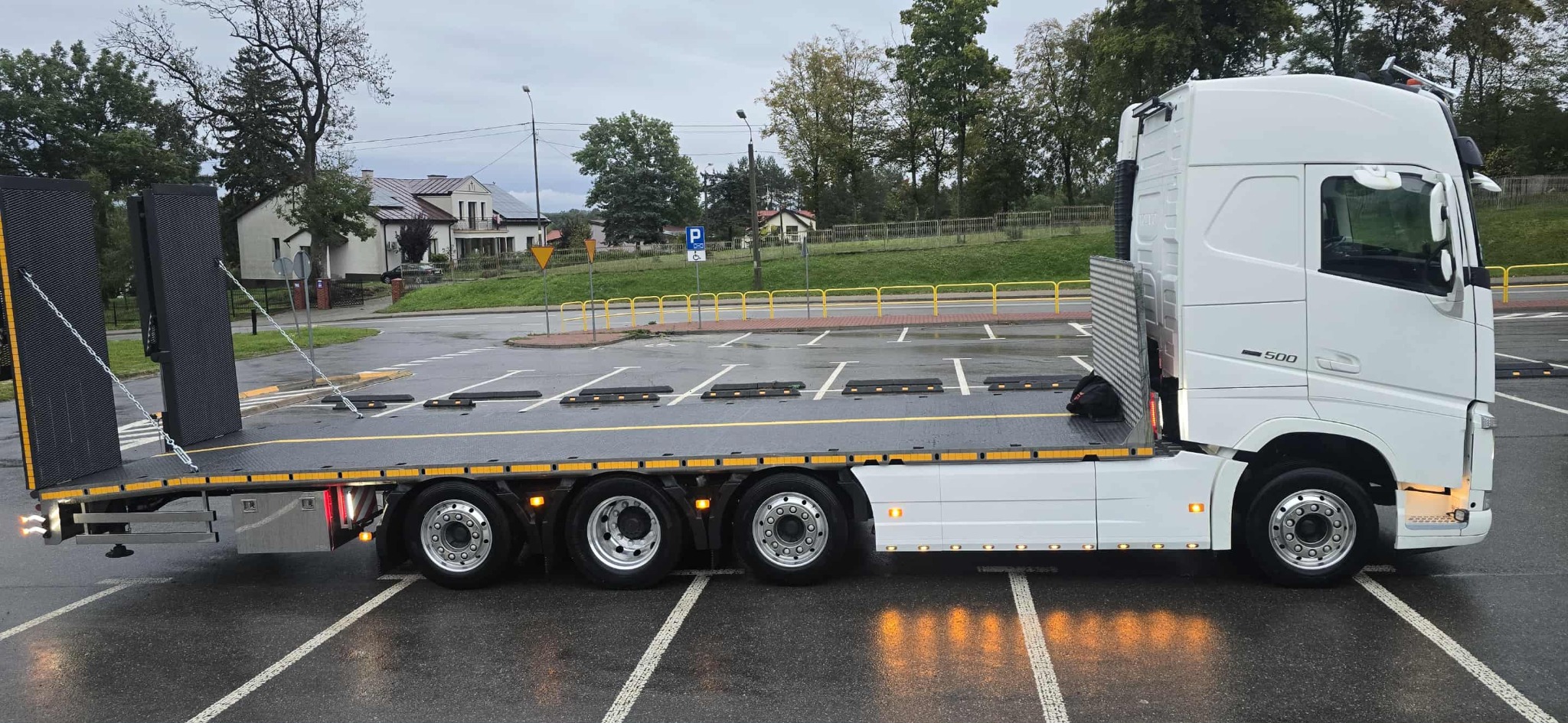 Biała ciężarówka z platformą transportową na parkingu, zabezpieczona łańcuchami. Widoczne detale konstrukcyjne i oświetlenie pojazdu. Tło stanowią budynki i drzewa.