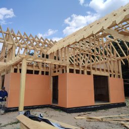 Dawid Salamon - Wznoszenie konstrukcji domu z drewna, widoczny szkielet dachu i ściany z panelami izolacyjnymi, pracownik w odzieży roboczej z wkrętarką stoi obok, na szczycie konstrukcji wieniec z gałęzi.