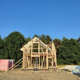 Dawid Salamon - Szkielet drewnianego domu w trakcie budowy, widoczne belki konstrukcyjne, drabina oparta o ścianę, materiały budowlane obok i pracownik w tle, otoczenie leśne.