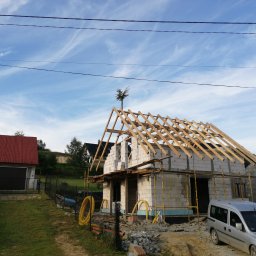 Dawid Salamon - Wznoszenie ścian z bloczków i konstrukcji dachu z drewna na budowie domu jednorodzinnego w Krynicy-Zdroju, widok z zewnątrz na częściowo ukończony budynek z szarym samochodem w tle.