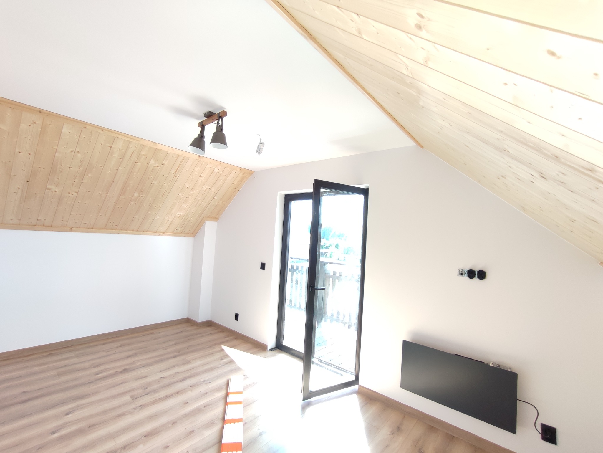 Poddasze z drewnianą boazerią na skosach, otwartymi drzwiami balkonowymi i grzejnikiem na ścianie.