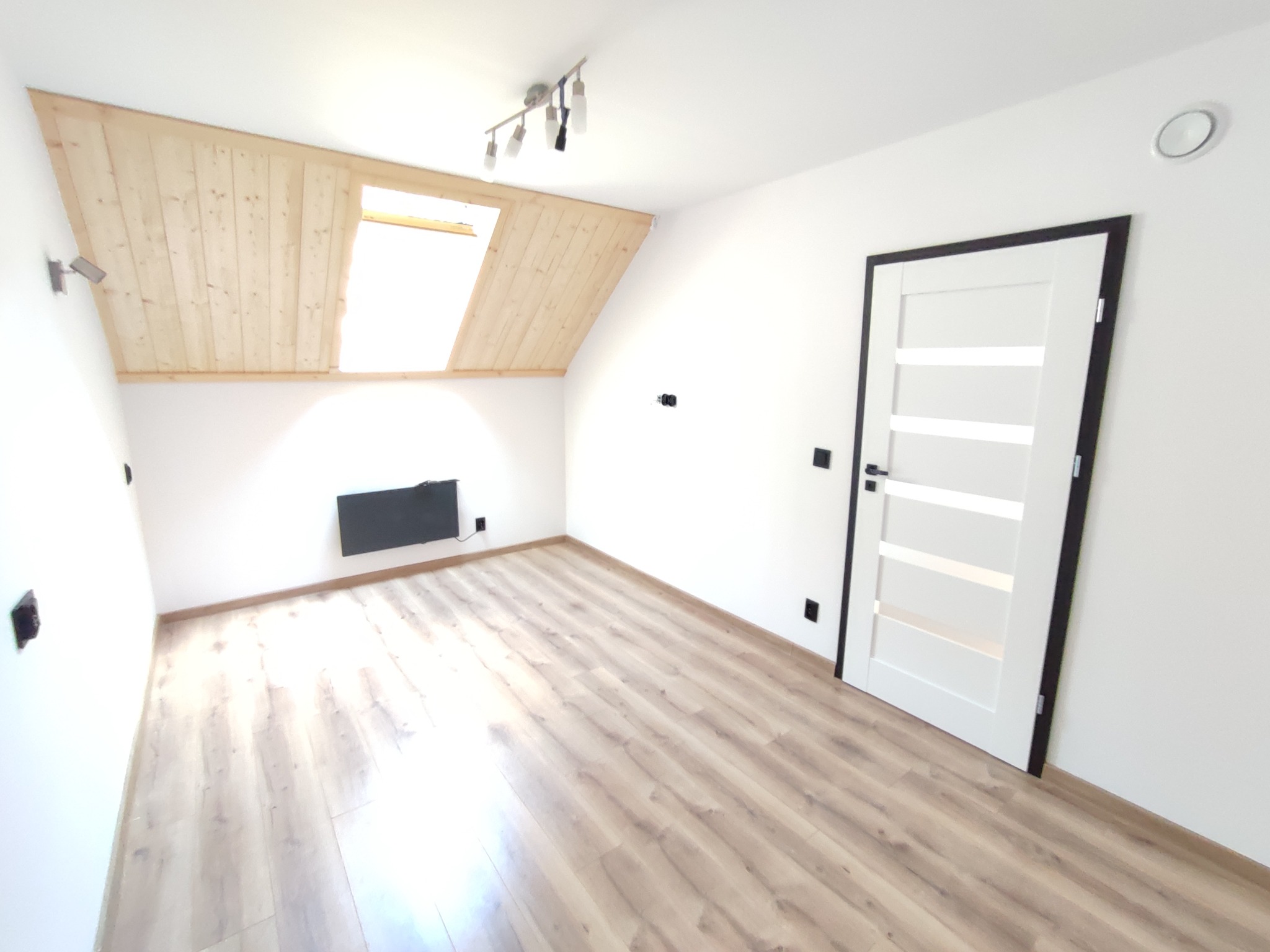 Poddasze z drewnianą boazerią na skosie sufitu, oknem dachowym i nową białą stolarką drzwiową z czarnymi ramami, panele podłogowe imitujące drewno, minimalistyczne oświetlenie