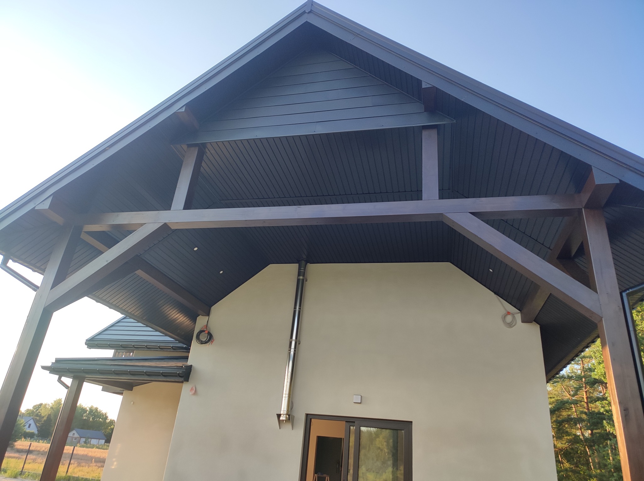 Nowoczesny dom z beżową elewacją i ciemnym drewnianym zadaszeniem wspartym na belkach, widoczny komin ze stali nierdzewnej i przesuwne drzwi balkonowe.