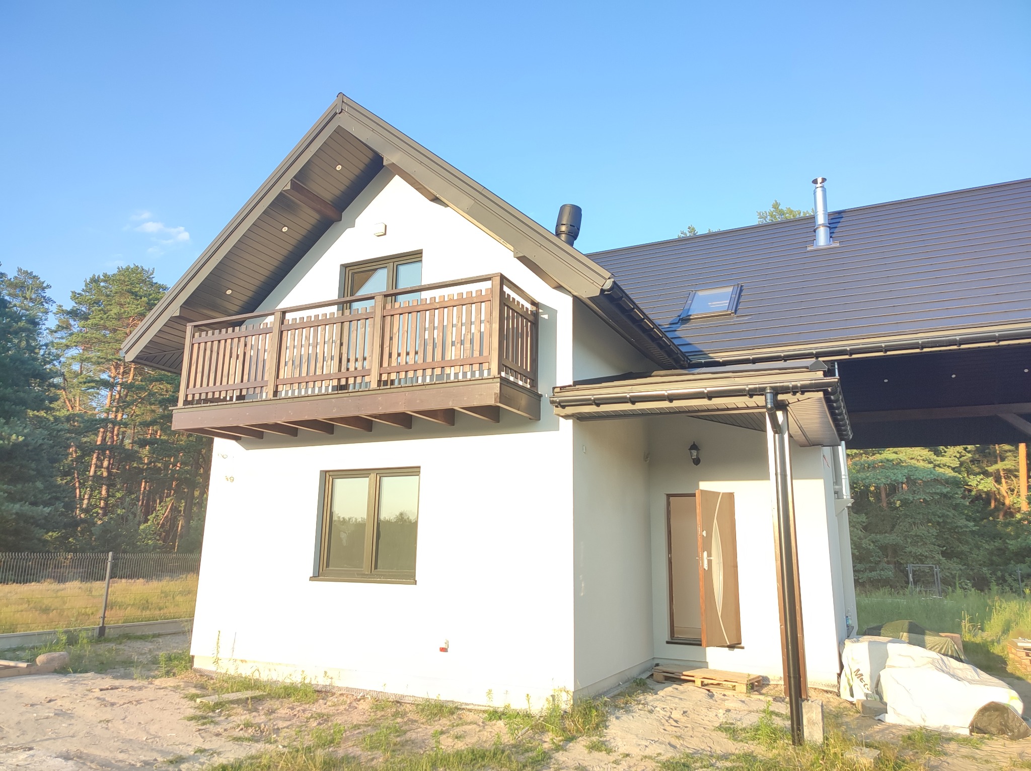 Nowo wybudowany dom z białymi ścianami, brązowym dachem i drewnianym balkonem, widok z zewnątrz w słoneczny dzień, teren wokół domu jeszcze niezagospodarowany.