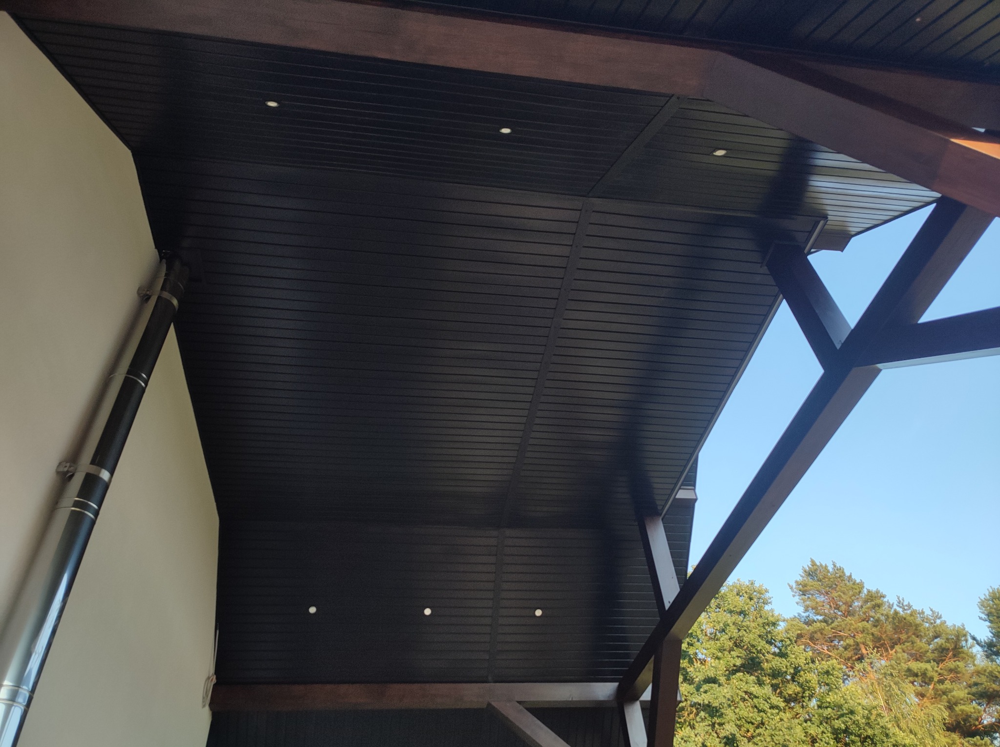 Zadaszenie tarasu z ciemnego drewna, widoczne od dołu, z elementami konstrukcyjnymi i fragmentem nieba w tle.