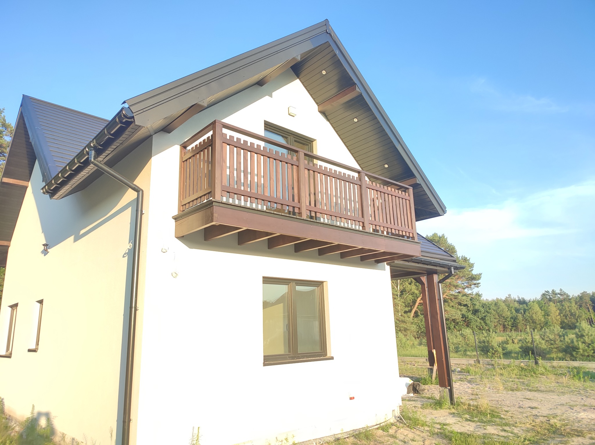 Nowoczesny dom murowany z balkonem z drewnianą balustradą, grafitowy dach, biała elewacja, widok z zewnątrz na tle błękitnego nieba i drzew.