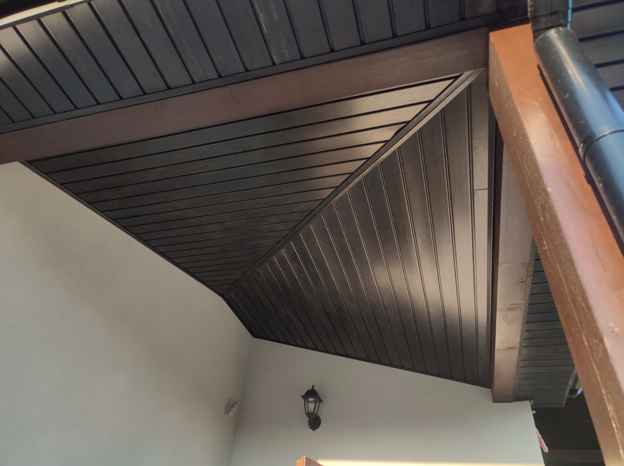 Podbitka dachowa z ciemnobrązowych paneli z widoczną lampą zewnętrzną na jasnej ścianie i brązowym słupem wspierającym.