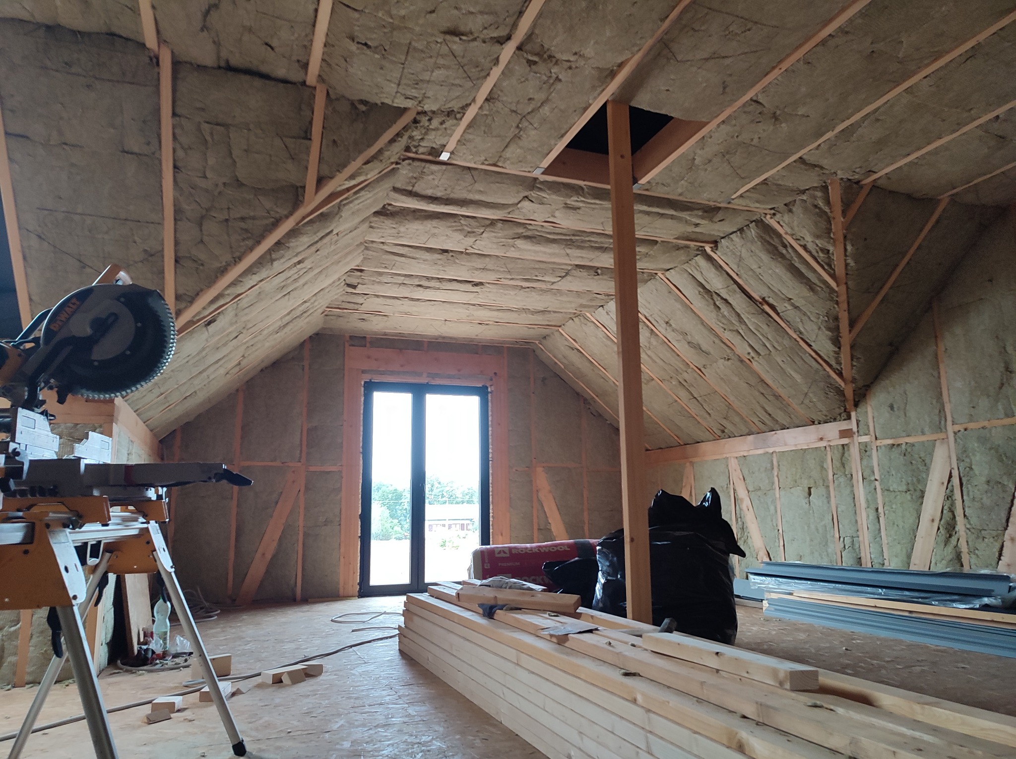 Poddasze w trakcie izolacji, widoczne wełna mineralna między krokwiami, przygotowane deski, piła ukośnica i okno dachowe.