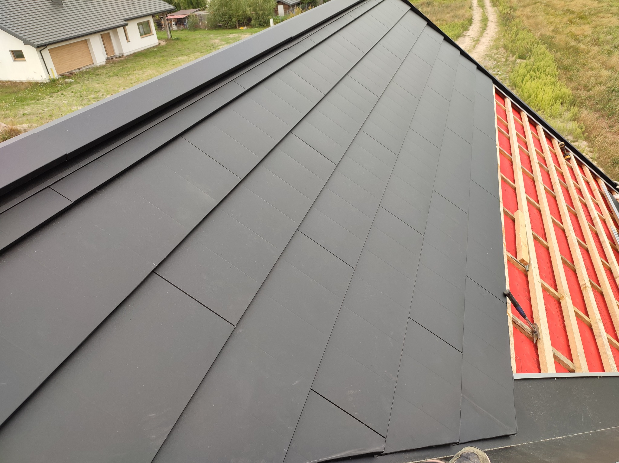 Fragment dachu z szarej blachy na rąbek stojący, z widoczną konstrukcją drewnianą i czerwoną membraną w niezadaszonej części, oraz fragmentem budynku w tle.