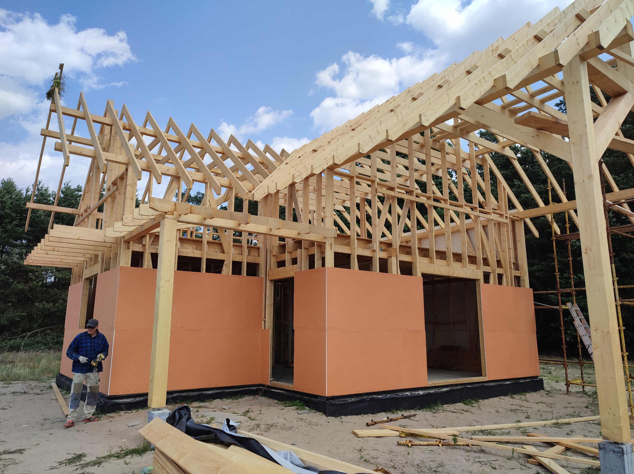 Wznoszenie konstrukcji domu z drewna, widoczny szkielet dachu i ściany z panelami izolacyjnymi, pracownik w odzieży roboczej z wkrętarką stoi obok, na szczycie konstrukcji wieniec z gałęzi.