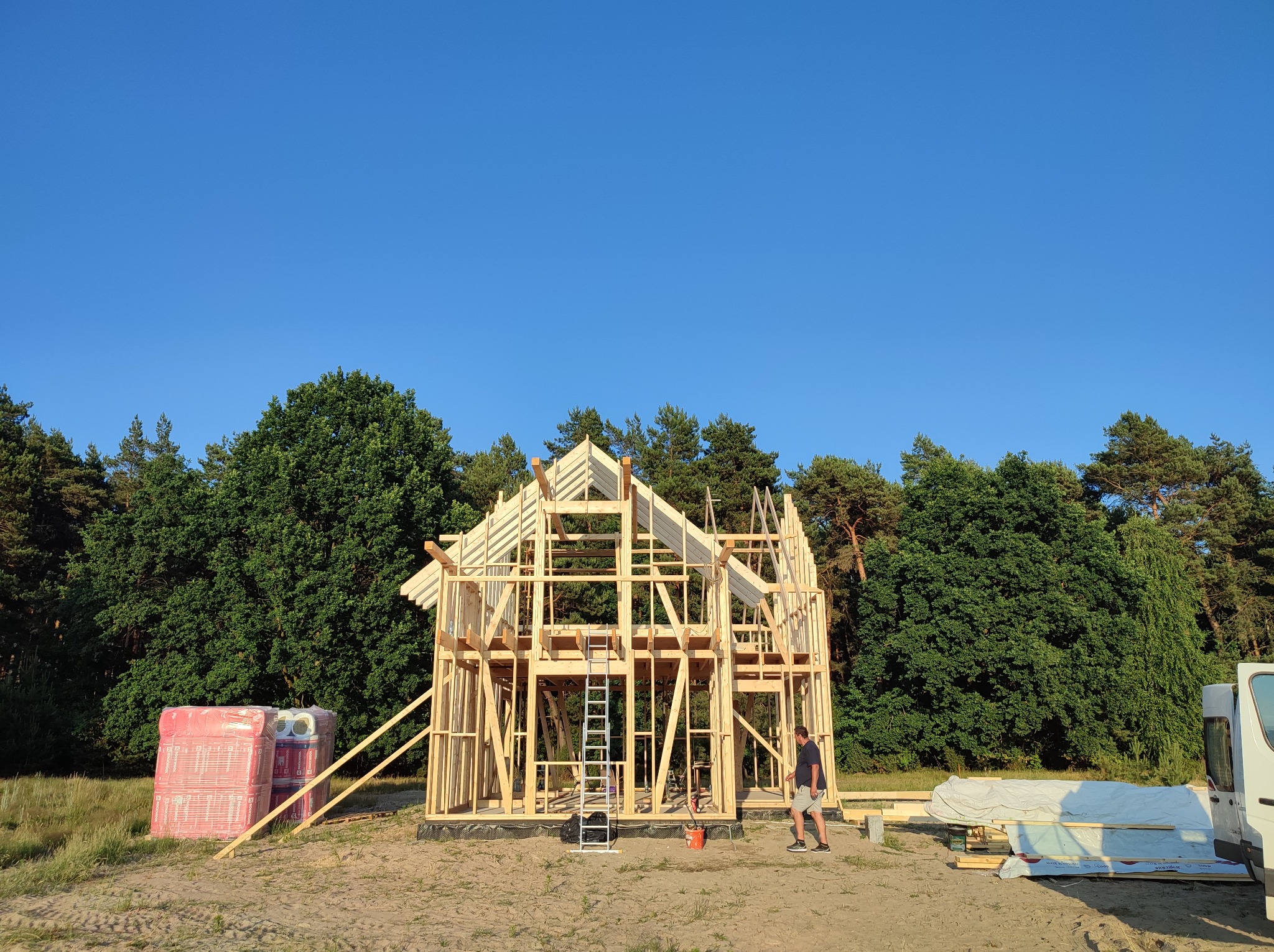 Szkielet drewnianego domu w trakcie budowy, widoczne belki konstrukcyjne, drabina oparta o ścianę, materiały budowlane obok i pracownik w tle, otoczenie leśne.