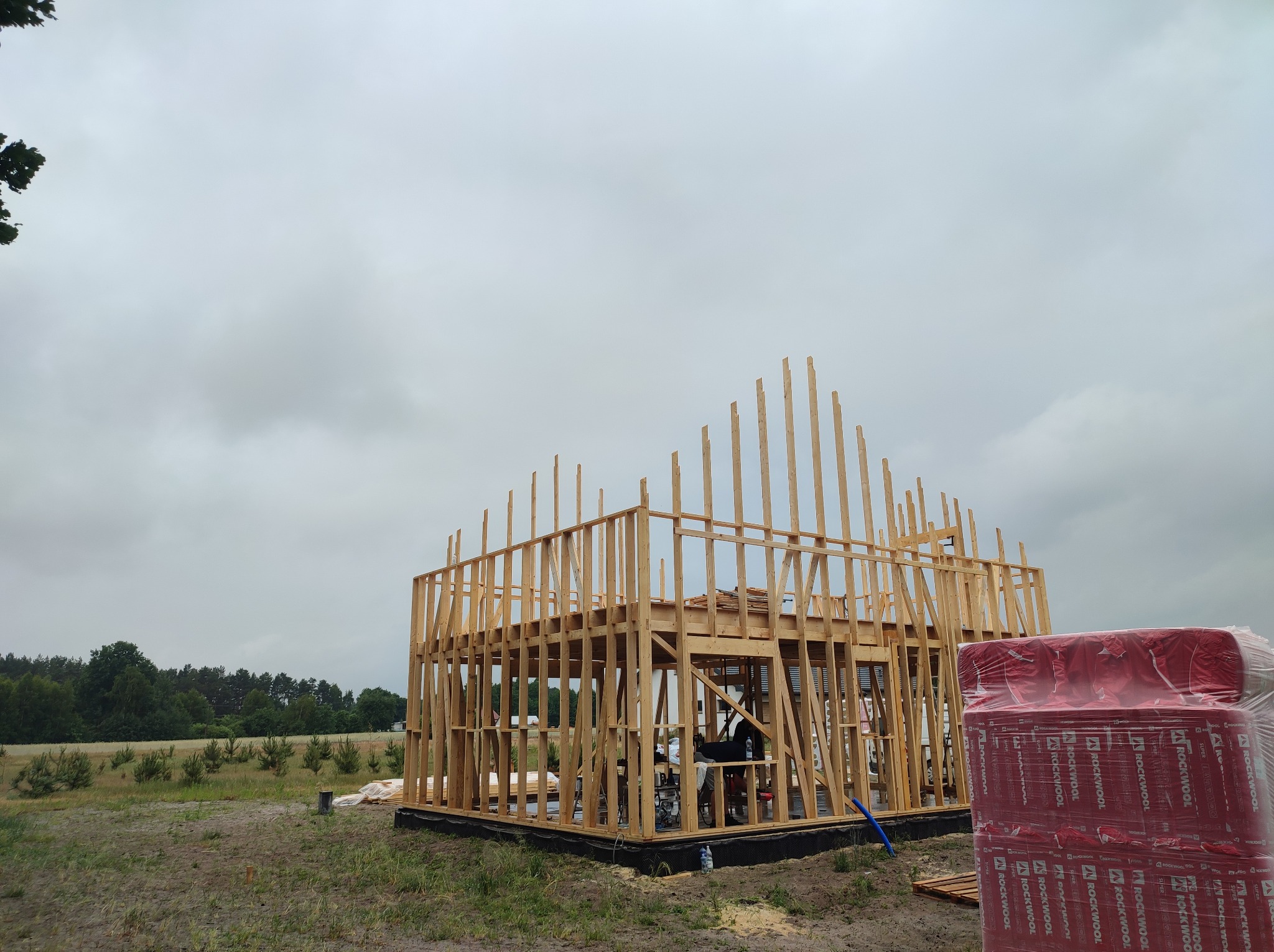 Konstrukcja szkieletowa domu w trakcie budowy, widoczne drewniane ramy ścian i stropu, na pierwszym planie zapakowane paczki wełny mineralnej Rockwool, pochmurne niebo w tle.