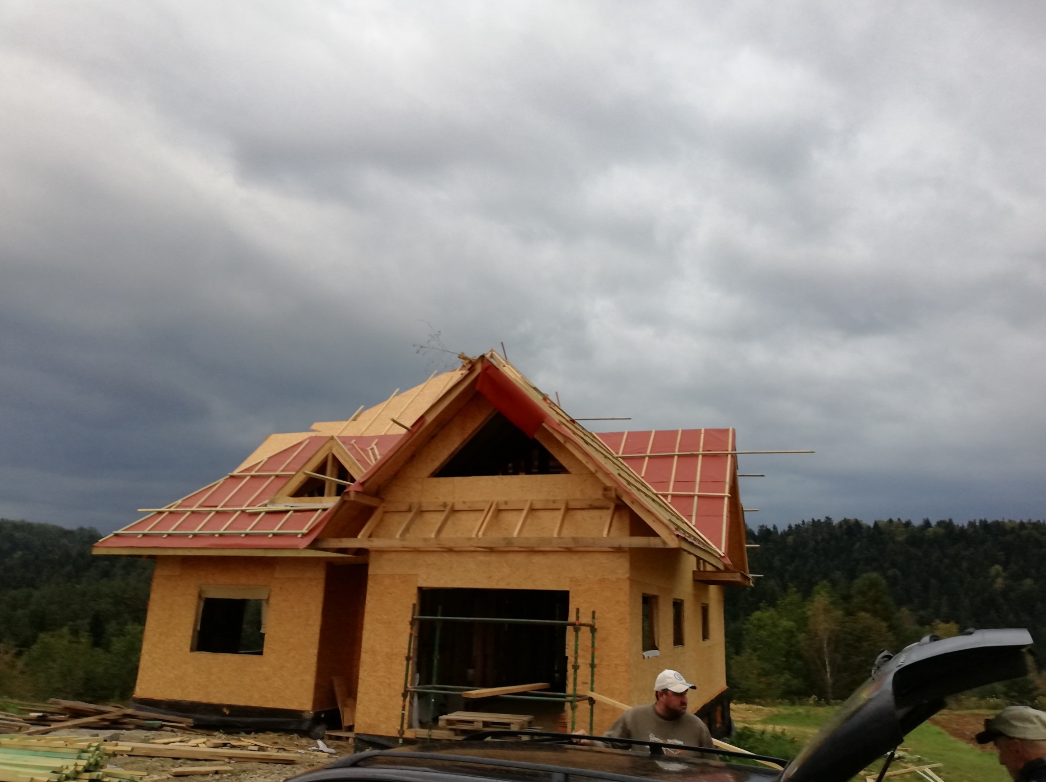 Wznoszenie konstrukcji domu z drewnianych elementów na tle pochmurnego nieba i zalesionego wzgórza, widoczne fragmenty czerwonego pokrycia dachowego i pracownicy przy samochodzie z otwartą maską.