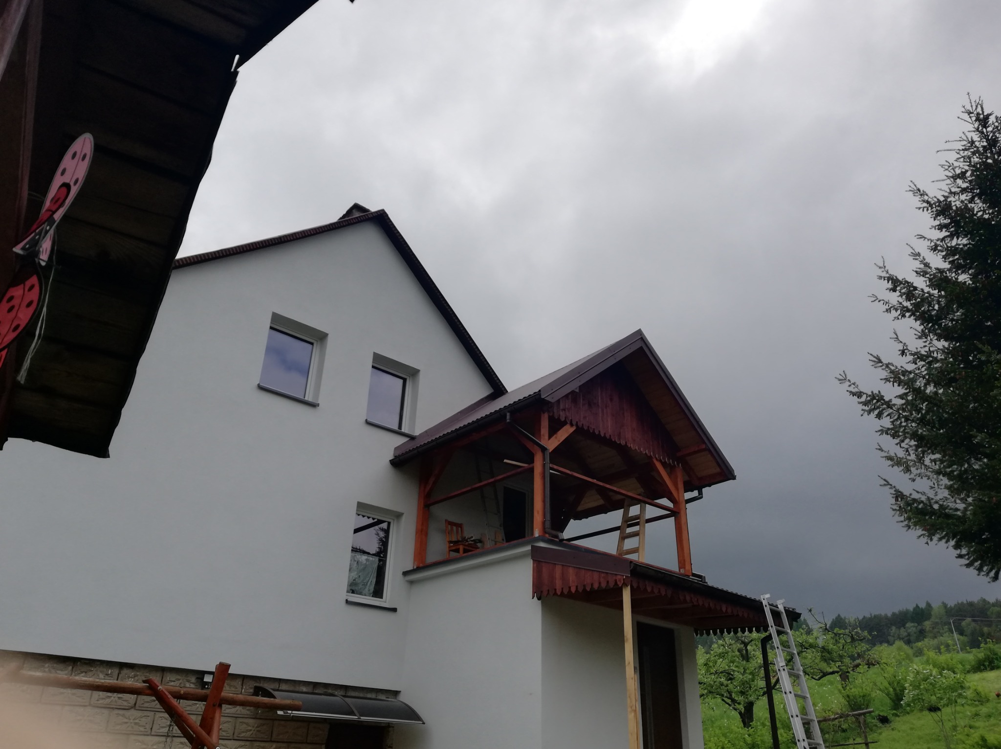 Biały dom z dobudowanym, drewnianym zadaszeniem balkonu, widok z dołu na pochmurne niebo, drabina oparta o ścianę, dekoracja w kształcie biedronki na zadaszeniu.