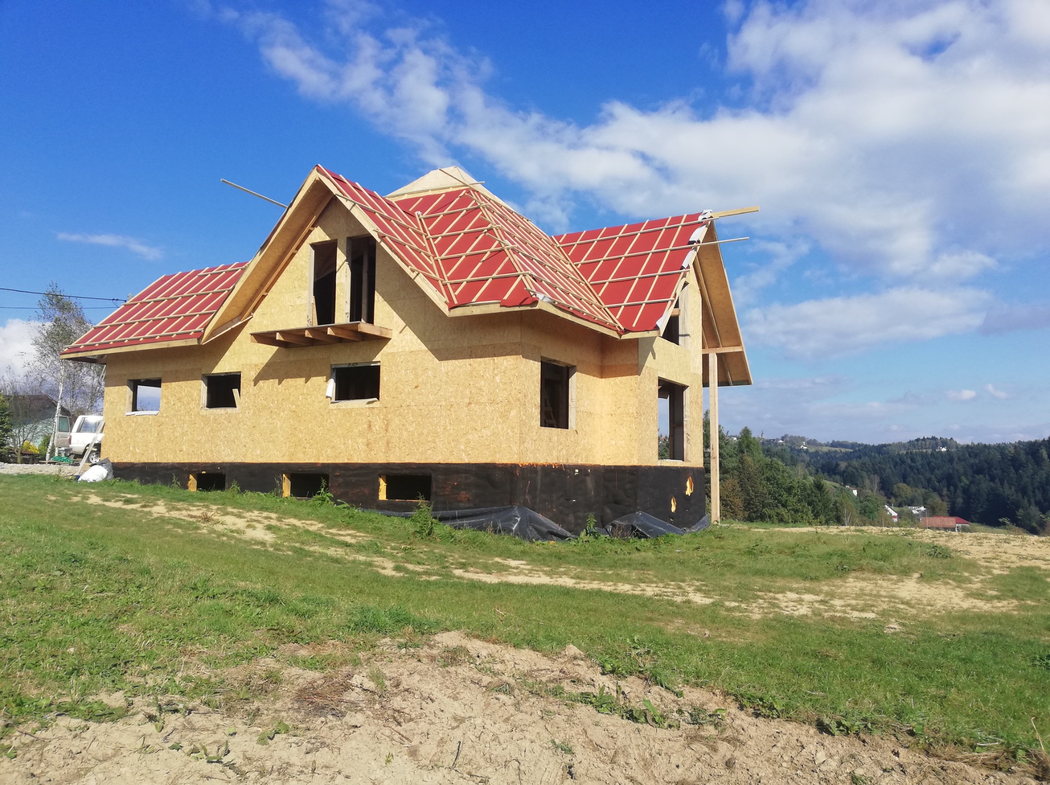 Surowy stan domu w konstrukcji szkieletowej z czerwonym pokryciem dachowym, widoczne czarne zabezpieczenie fundamentów, na tle zielonego wzgórza i błękitnego nieba z chmurami.