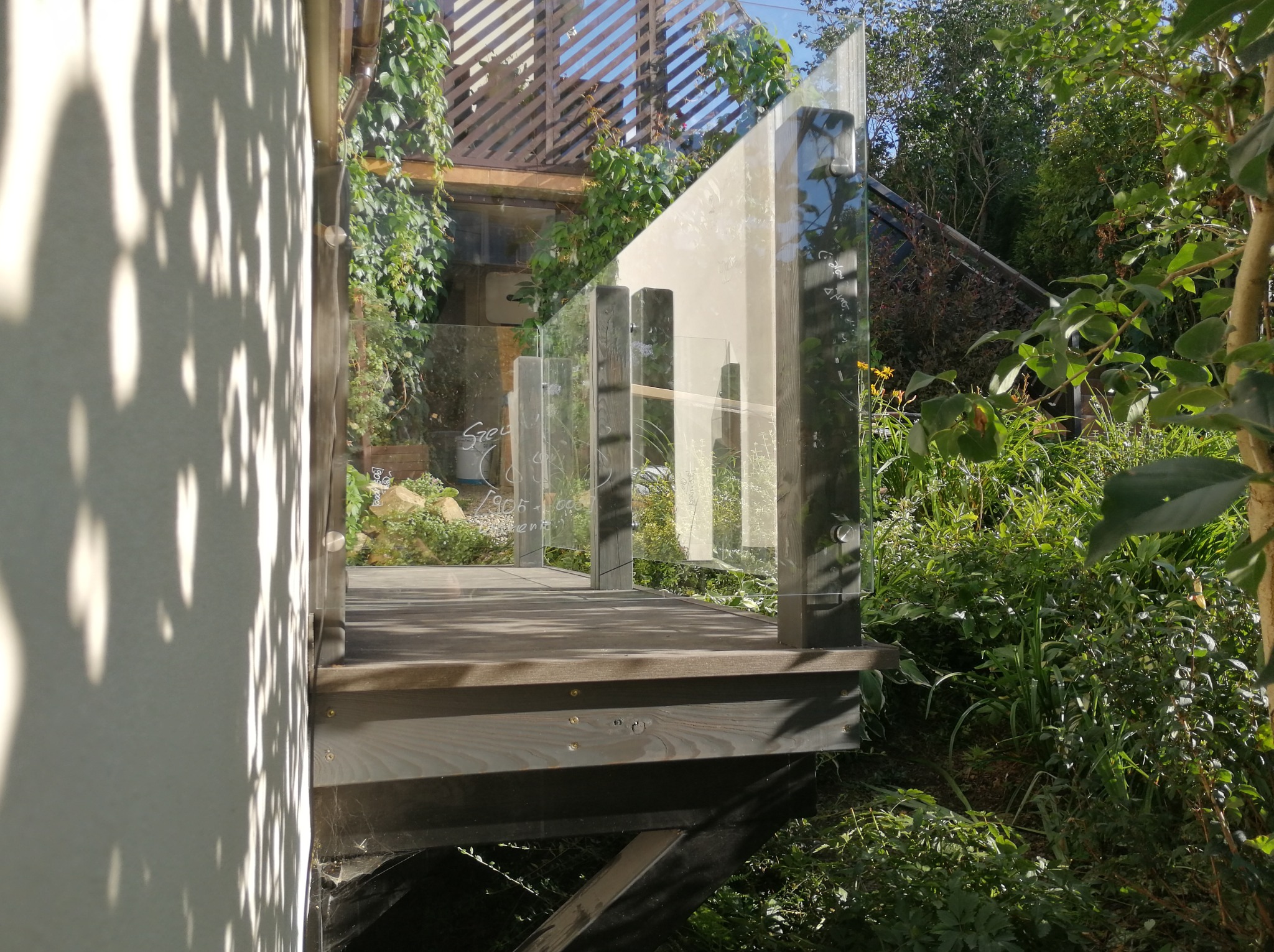 Drewniany taras z nowoczesną szklaną balustradą, otoczony bujną zielenią ogrodu, widoczne refleksy światła na szkle i cień liści na ścianie budynku.