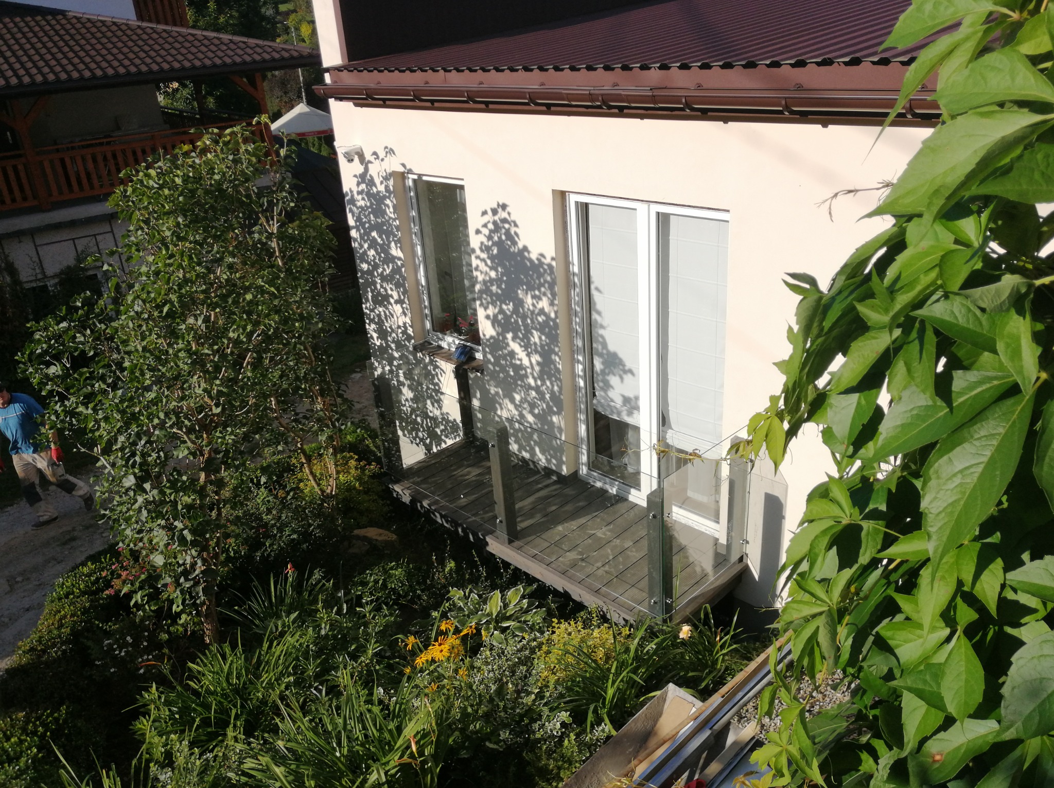 Widok z góry na fragment domu z balkonem z szklaną balustradą, otoczony zielenią i częściowo zasłonięty przez bujną roślinność, z widocznym fragmentem dachu i osoby w oddali.