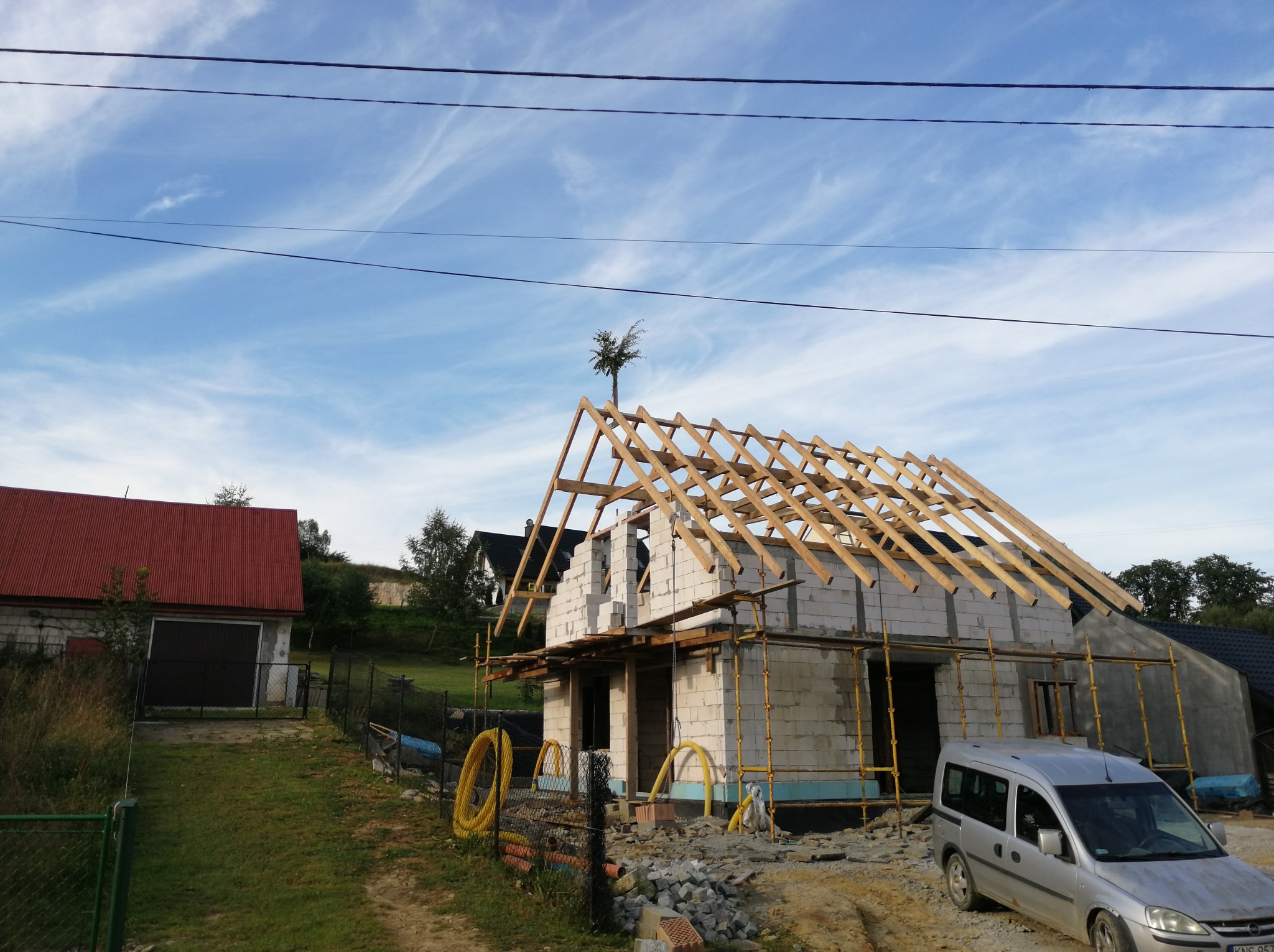 Wznoszenie ścian z bloczków i konstrukcji dachu z drewna na budowie domu jednorodzinnego w Krynicy-Zdroju, widok z zewnątrz na częściowo ukończony budynek z szarym samochodem w tle.