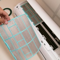 DAMIAN MILCZEK DM-AQUA USŁUGI INSTALACYJNE I WENTYLACJA - Brudny, wyjmowany filtr powietrza klimatyzatora z turkusową ramką, trzymany w dłoni, obok otwartej obudowy klimatyzatora.