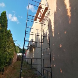 DAMIAN MILCZEK DM-AQUA USŁUGI INSTALACYJNE I WENTYLACJA - Pracownik na rusztowaniu podczas montażu klimatyzacji zewnętrznej na elewacji budynku, widoczne przewody i jednostka zewnętrzna.