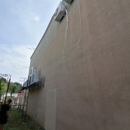 DAMIAN MILCZEK DM-AQUA USŁUGI INSTALACYJNE I WENTYLACJA - Montaż klimatyzacji na elewacji budynku, widoczne jednostki zewnętrzne i przewody, drabina rusztowaniowa z pracownikiem w tle.