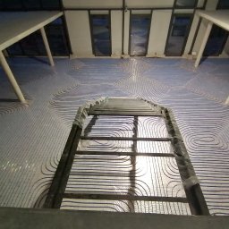 DAMIAN MILCZEK DM-AQUA USŁUGI INSTALACYJNE I WENTYLACJA - Ułożone rury ogrzewania podłogowego w jasnym pomieszczeniu, widoczne z góry przez metalową konstrukcję. Równoległe pętle rur tworzą wzór na podłodze.
