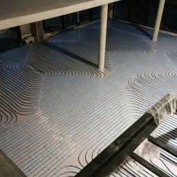 DAMIAN MILCZEK DM-AQUA USŁUGI INSTALACYJNE I WENTYLACJA - Ułożone, niebieskie rury ogrzewania podłogowego na siatce, widoczne słupy konstrukcyjne. Ujęcie z góry, w tle okna. Instalacja przed wylaniem posadzki.