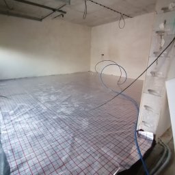 DAMIAN MILCZEK DM-AQUA USŁUGI INSTALACYJNE I WENTYLACJA - Rozłożona folia grzewcza z czerwonym rastrem na podłodze w surowym wnętrzu. Widoczne rury instalacji. Etap prac przed wylaniem posadzki.