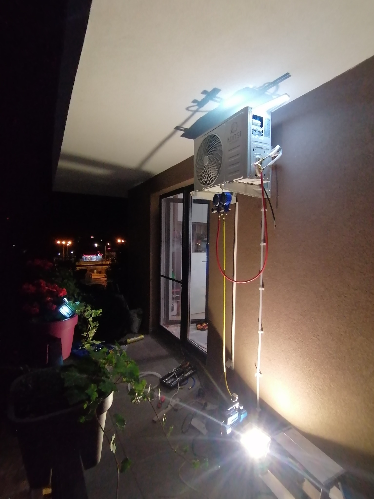 Montaż klimatyzatora typu split na balkonie w warunkach nocnych, widoczne przewody, manometry i lampa oświetlająca miejsce pracy.