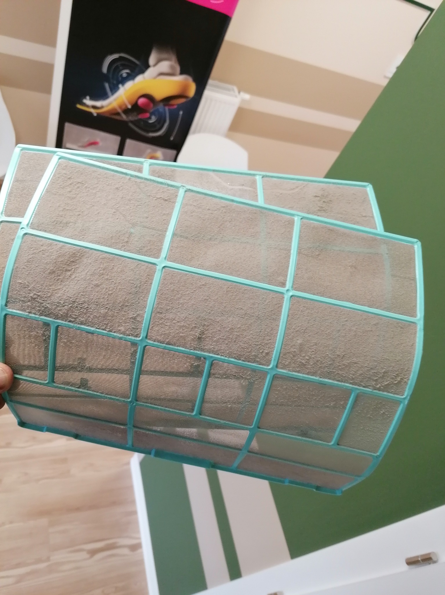 Dwa bardzo brudne filtry powietrza klimatyzacji z niebieską ramką, trzymane w dłoni, na tle ściany i plakatu reklamowego.