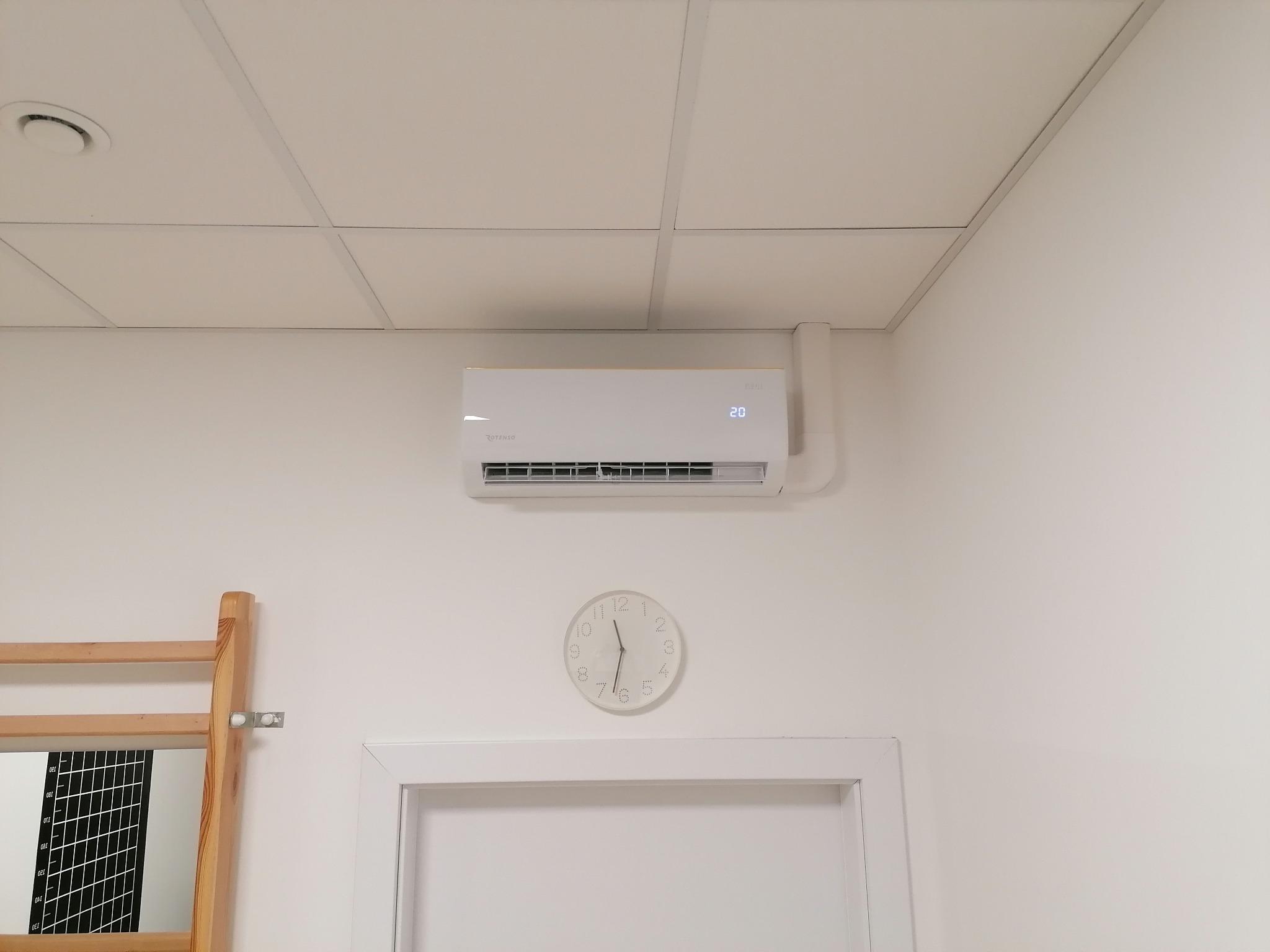 Biały klimatyzator ścienny marki Rotenso z wyświetlaczem temperatury 20 stopni Celsjusza, zamontowany nad białymi drzwiami i okrągłym zegarem, widoczna drabinka gimnastyczna z boku.