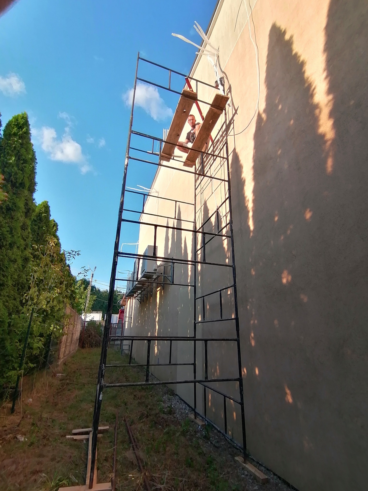 Pracownik na rusztowaniu podczas montażu klimatyzacji zewnętrznej na elewacji budynku, widoczne przewody i jednostka zewnętrzna.