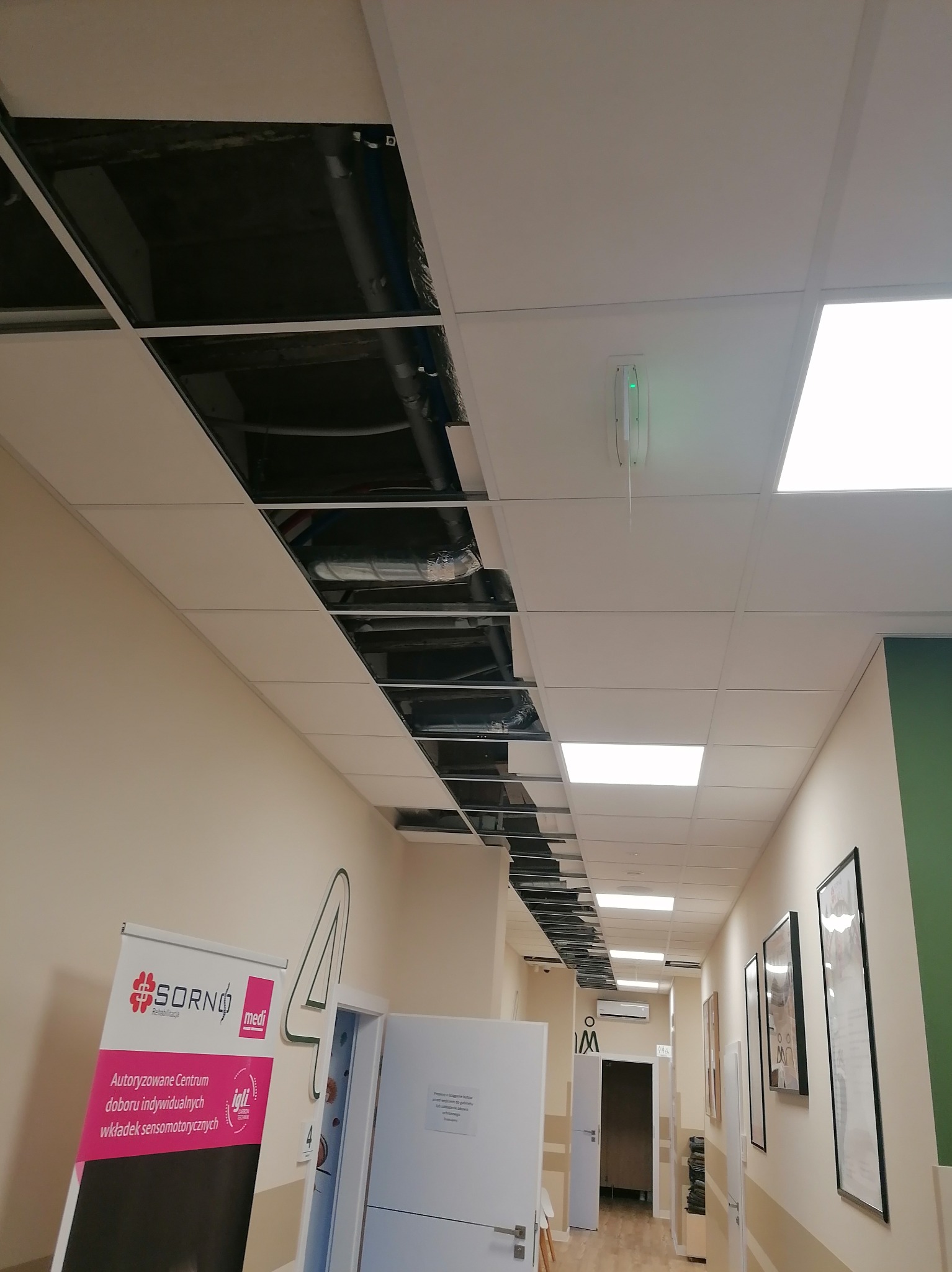 Sufit podwieszany z otwartymi panelami ukazującymi instalacje wentylacyjne i rury, widok z korytarza w budynku komercyjnym.