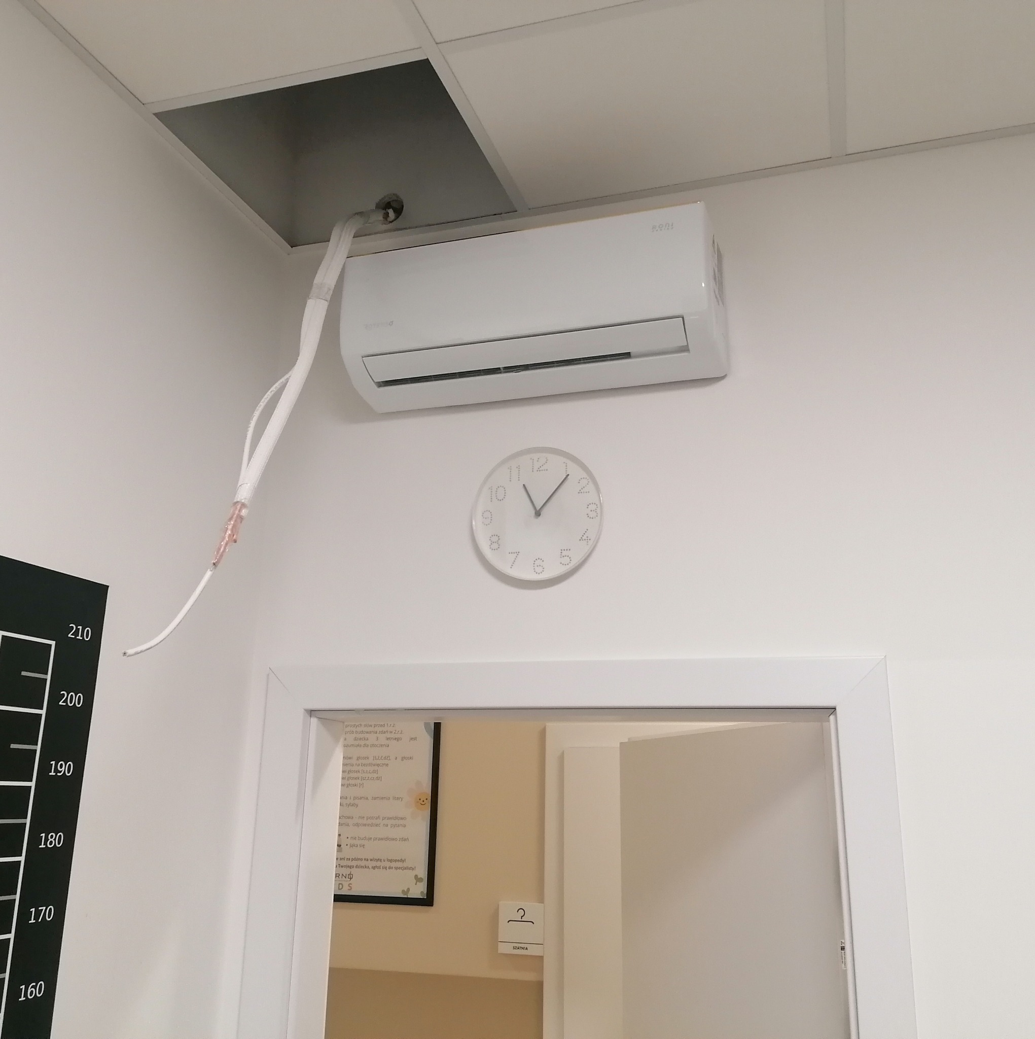 Biały klimatyzator ścienny zamontowany w pomieszczeniu z odkrytą przestrzenią w suficie podwieszanym, widoczne przewody instalacyjne, zegar na ścianie, obok drzwi i tablica ze skalą wzrostu.