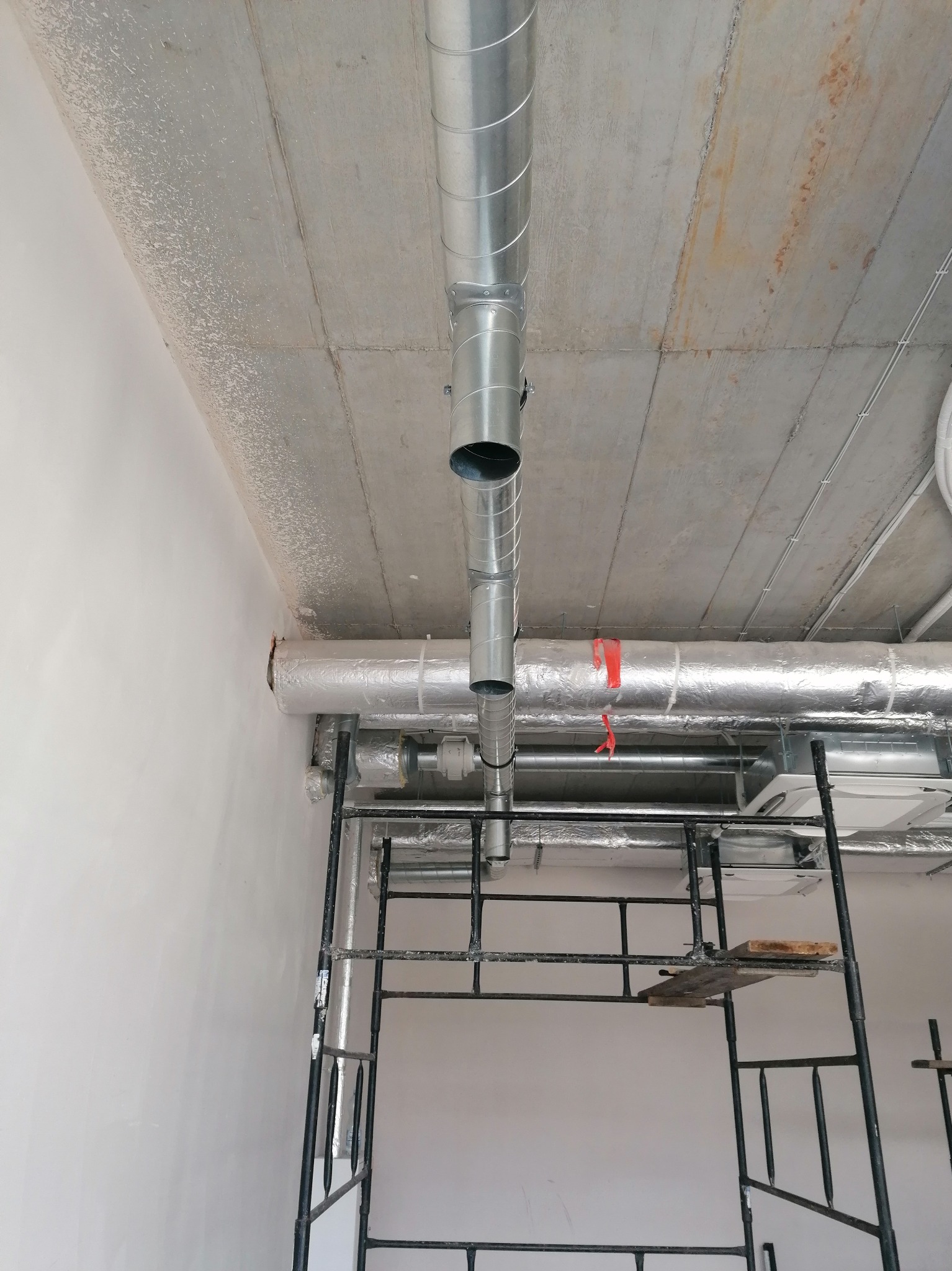 Montaż spiralnych rur wentylacyjnych z blachy ocynkowanej pod surowym betonowym sufitem, widoczne rusztowanie i klimatyzator w tle.