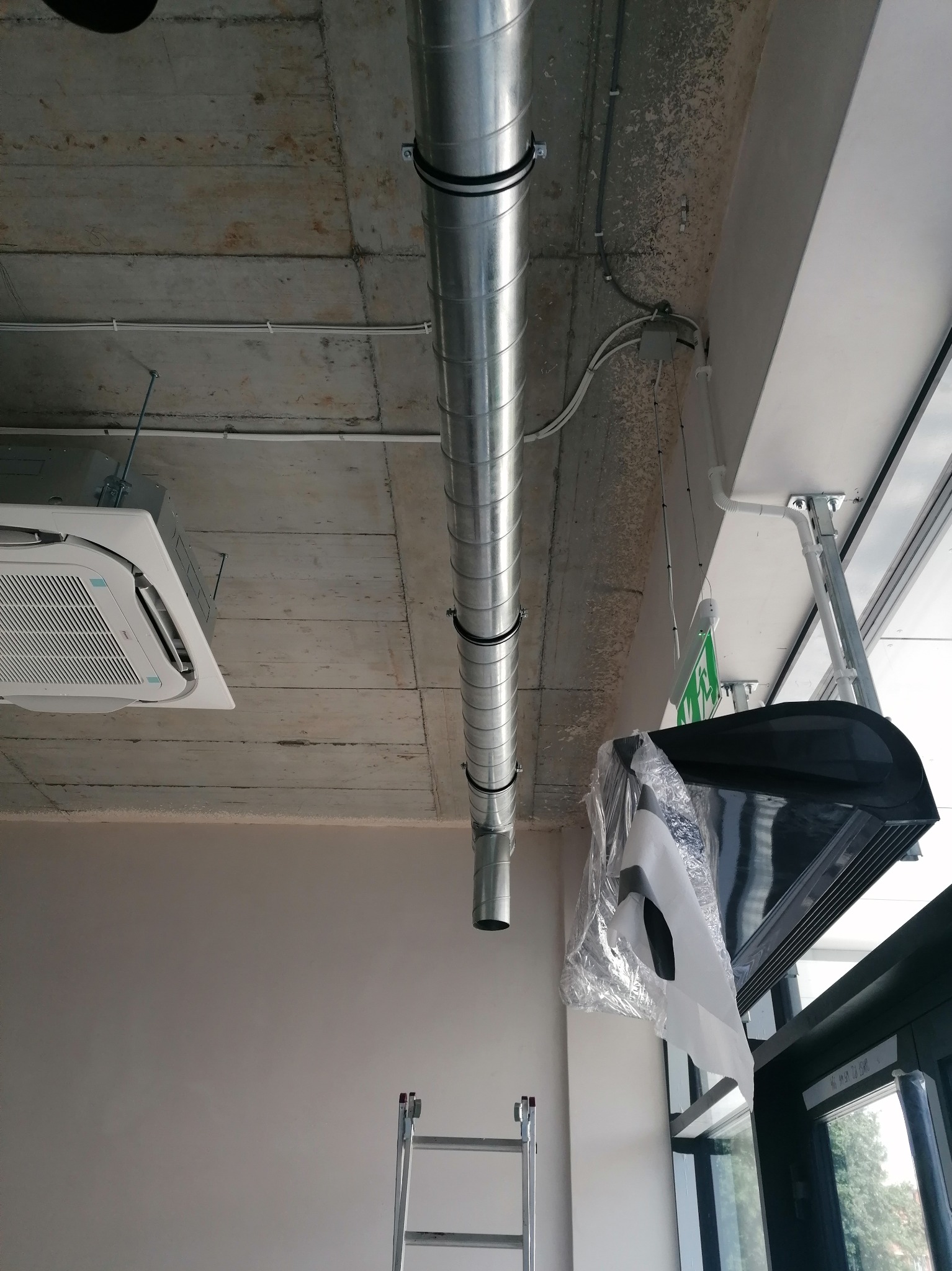 Montaż klimatyzacji kasetonowej z widocznym systemem kanałów wentylacyjnych pod surowym betonowym sufitem, drabina w tle.