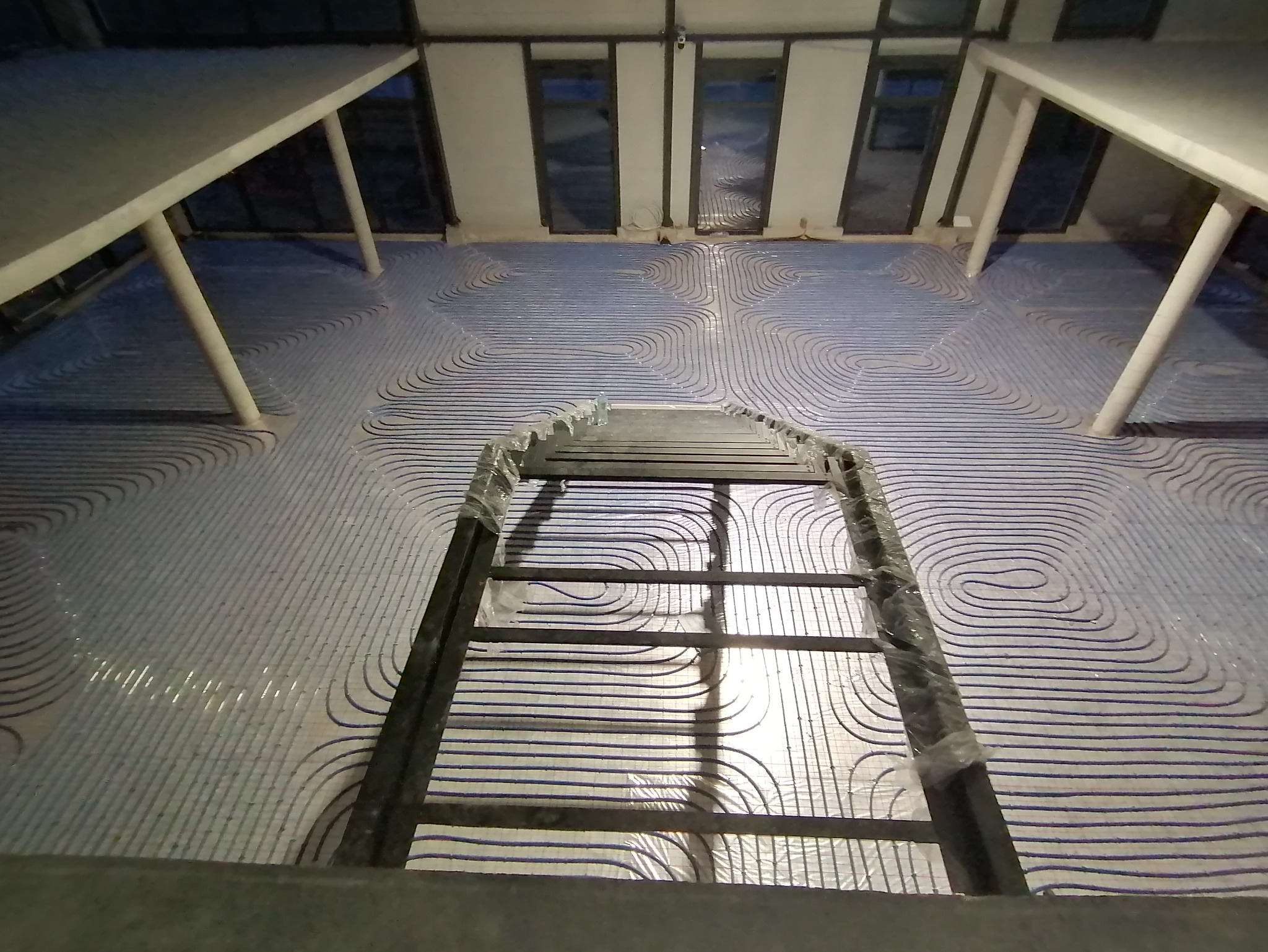 Ułożone rury ogrzewania podłogowego w jasnym pomieszczeniu, widoczne z góry przez metalową konstrukcję. Równoległe pętle rur tworzą wzór na podłodze.