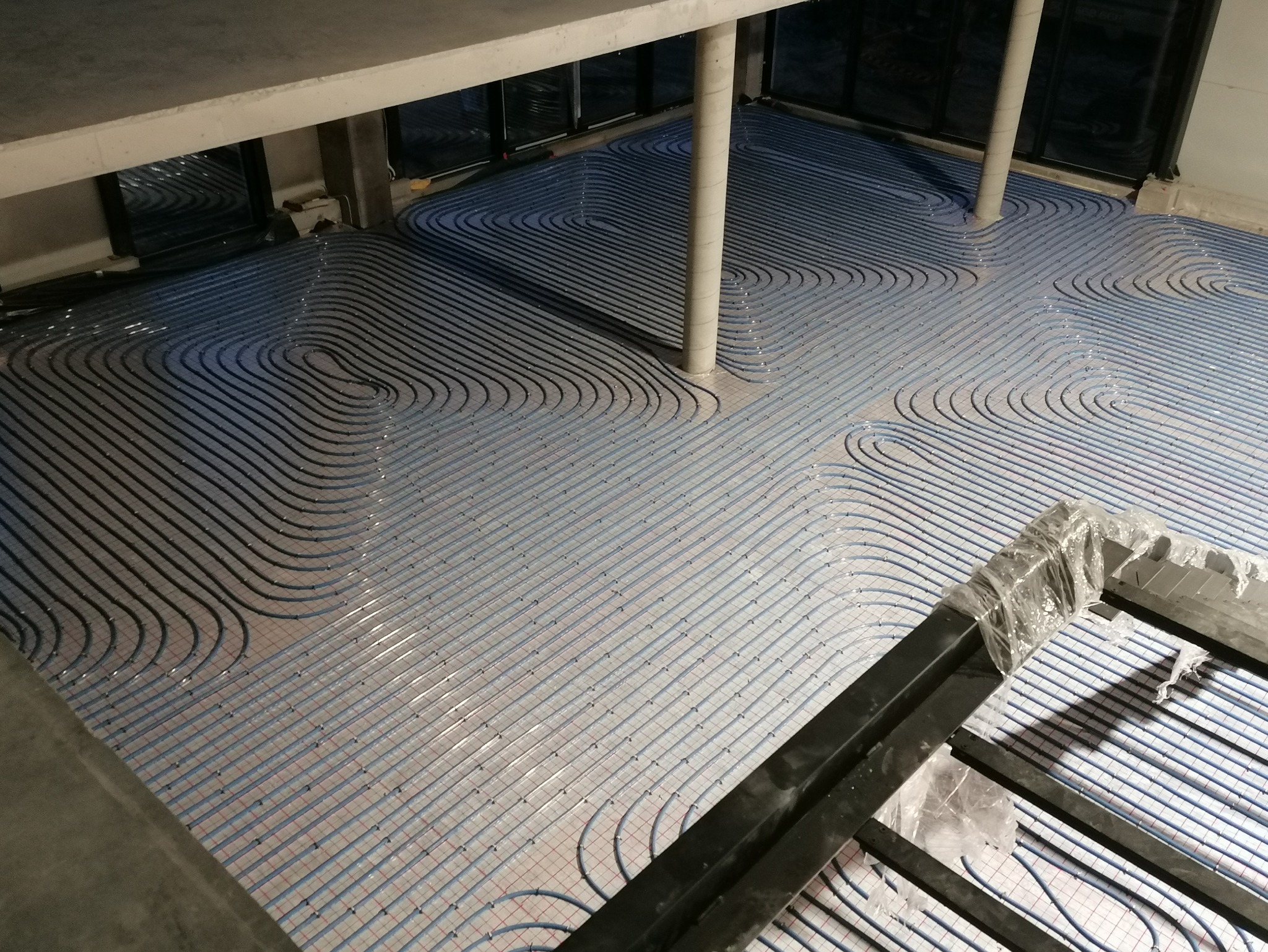 Ułożone, niebieskie rury ogrzewania podłogowego na siatce, widoczne słupy konstrukcyjne. Ujęcie z góry, w tle okna. Instalacja przed wylaniem posadzki.
