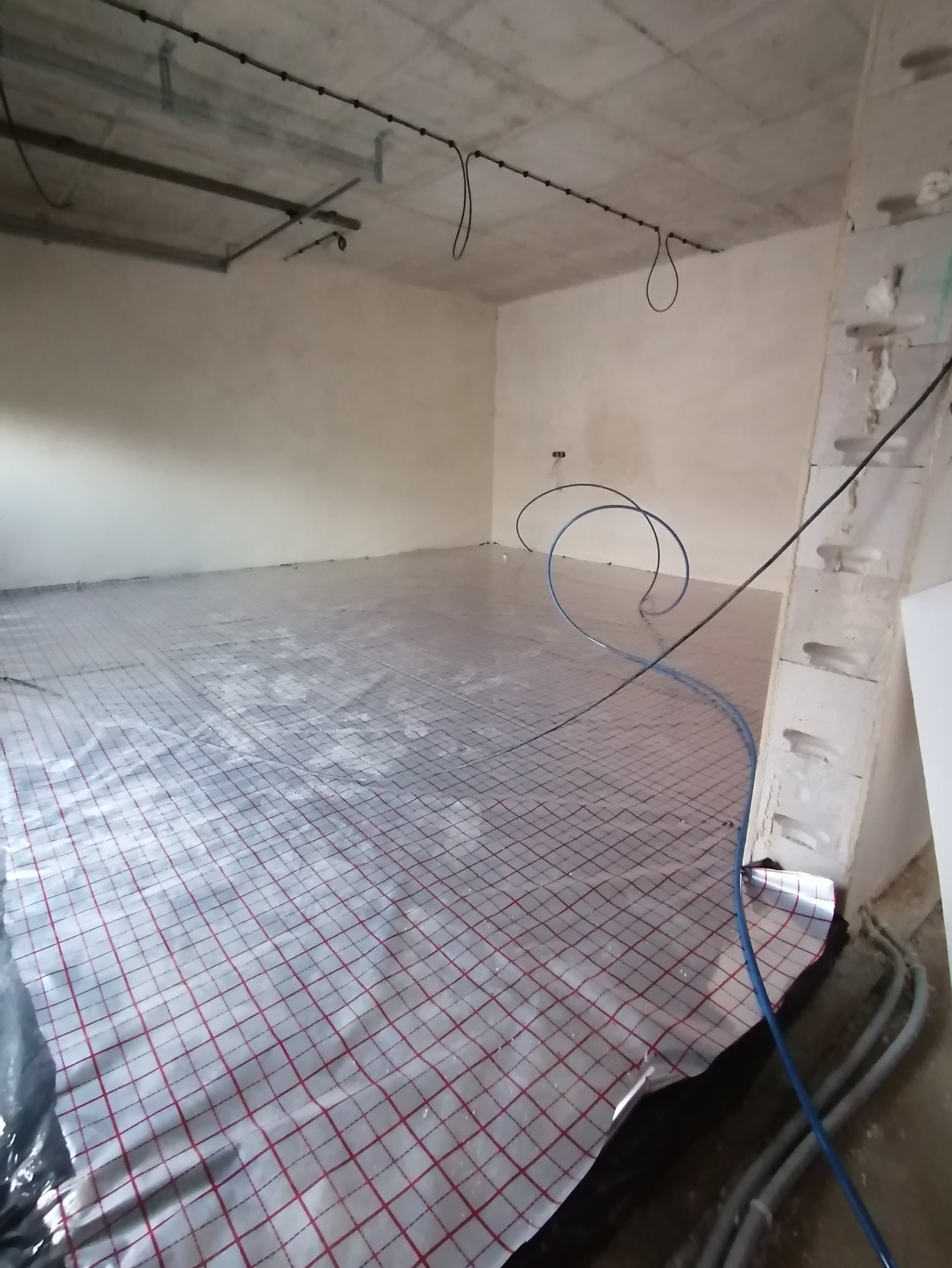 Rozłożona folia grzewcza z czerwonym rastrem na podłodze w surowym wnętrzu. Widoczne rury instalacji. Etap prac przed wylaniem posadzki.