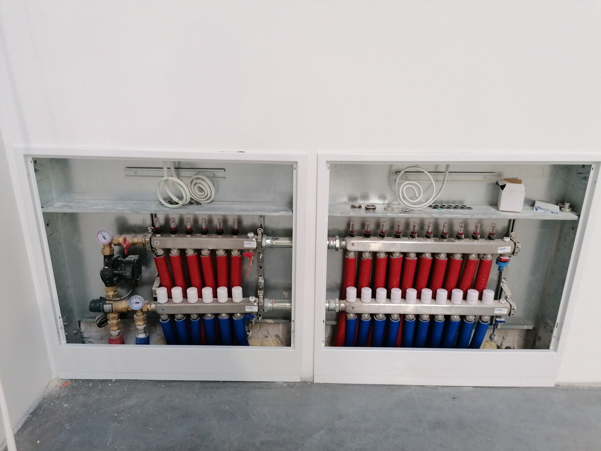 Dwa rozdzielacze ogrzewania podłogowego w metalowych szafkach, z rurami w kolorach czerwonym i niebieskim, zamontowane przy białej ścianie i szarej podłodze.