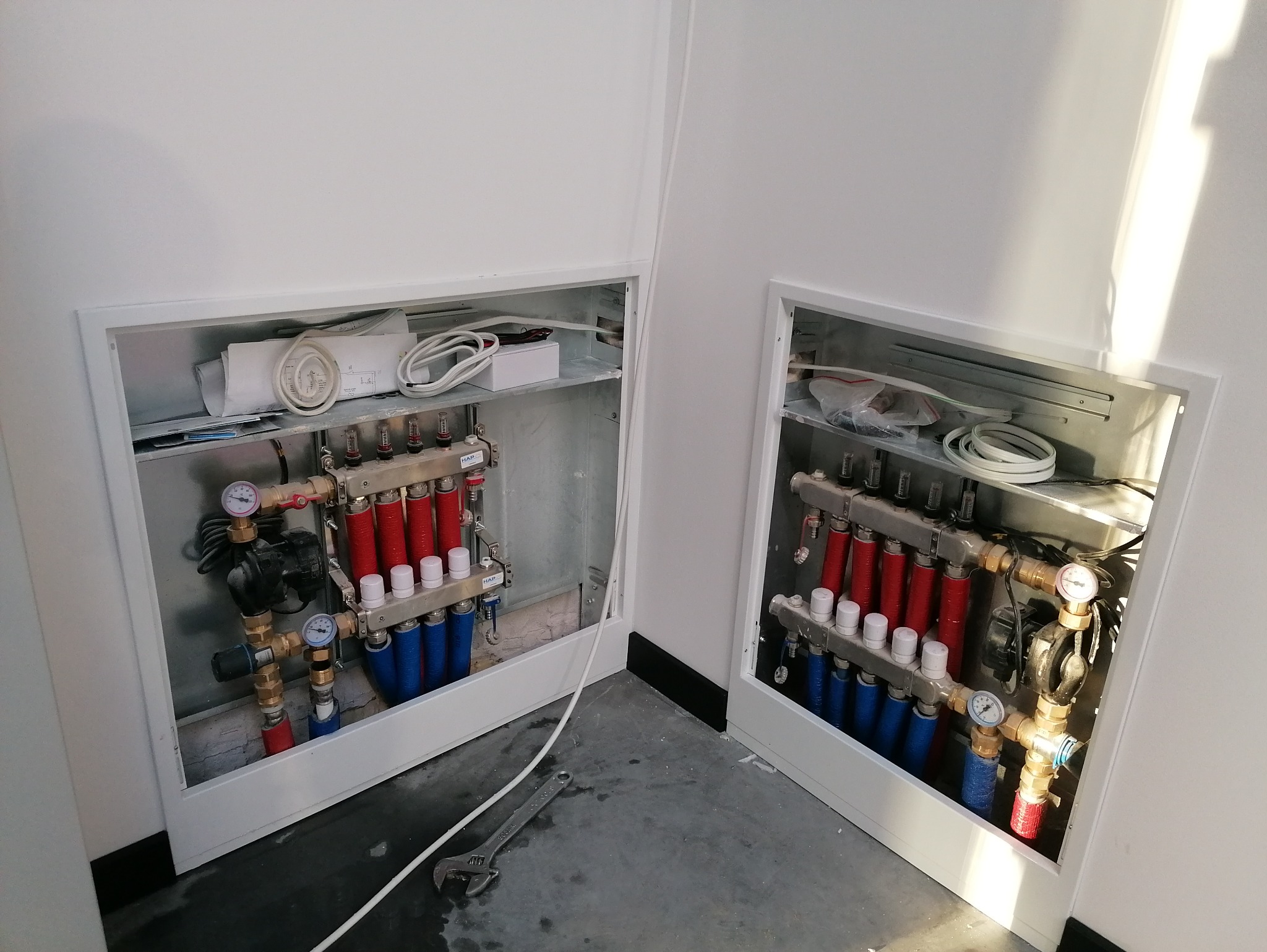 Dwa kolektory podłogowe w białych wnękach, z rurami czerwonymi i niebieskimi. Na podłodze leży klucz nastawny. Widoczne manometry i przewody.