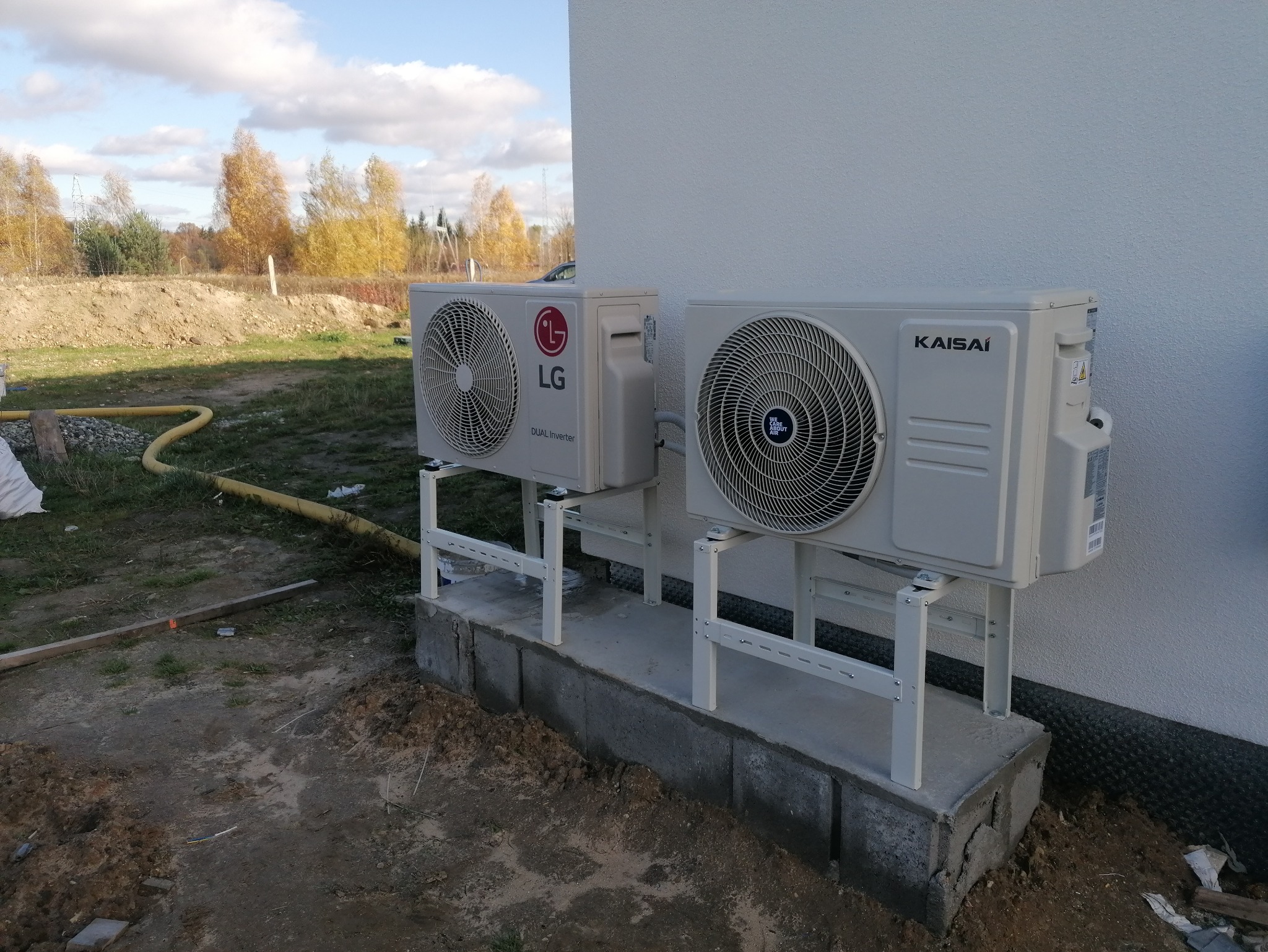 Dwa klimatyzatory (LG i Kaisai) zamontowane na zewnątrz budynku na betonowym fundamencie i metalowych wspornikach. Widoczne elementy instalacji i otoczenie budynku.