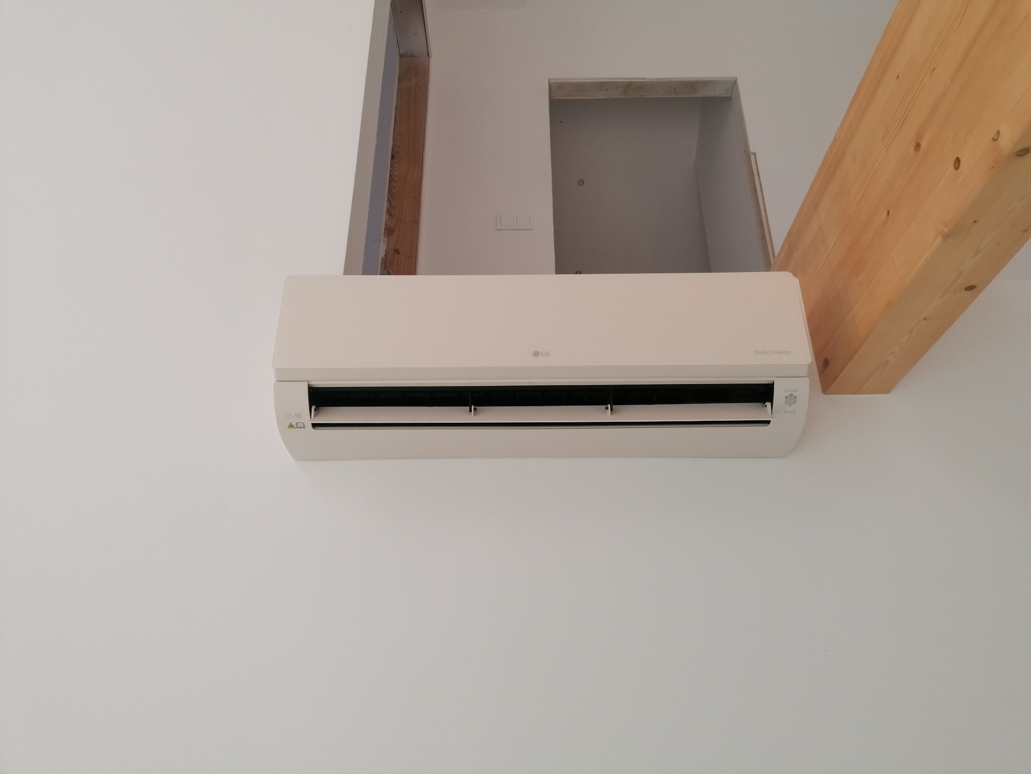 Zamontowany klimatyzator LG Dual Inverter na białej ścianie, widoczny fragment drewnianej belki stropowej i otwór w ścianie. Minimalistyczny design, nowoczesne wnętrze.
