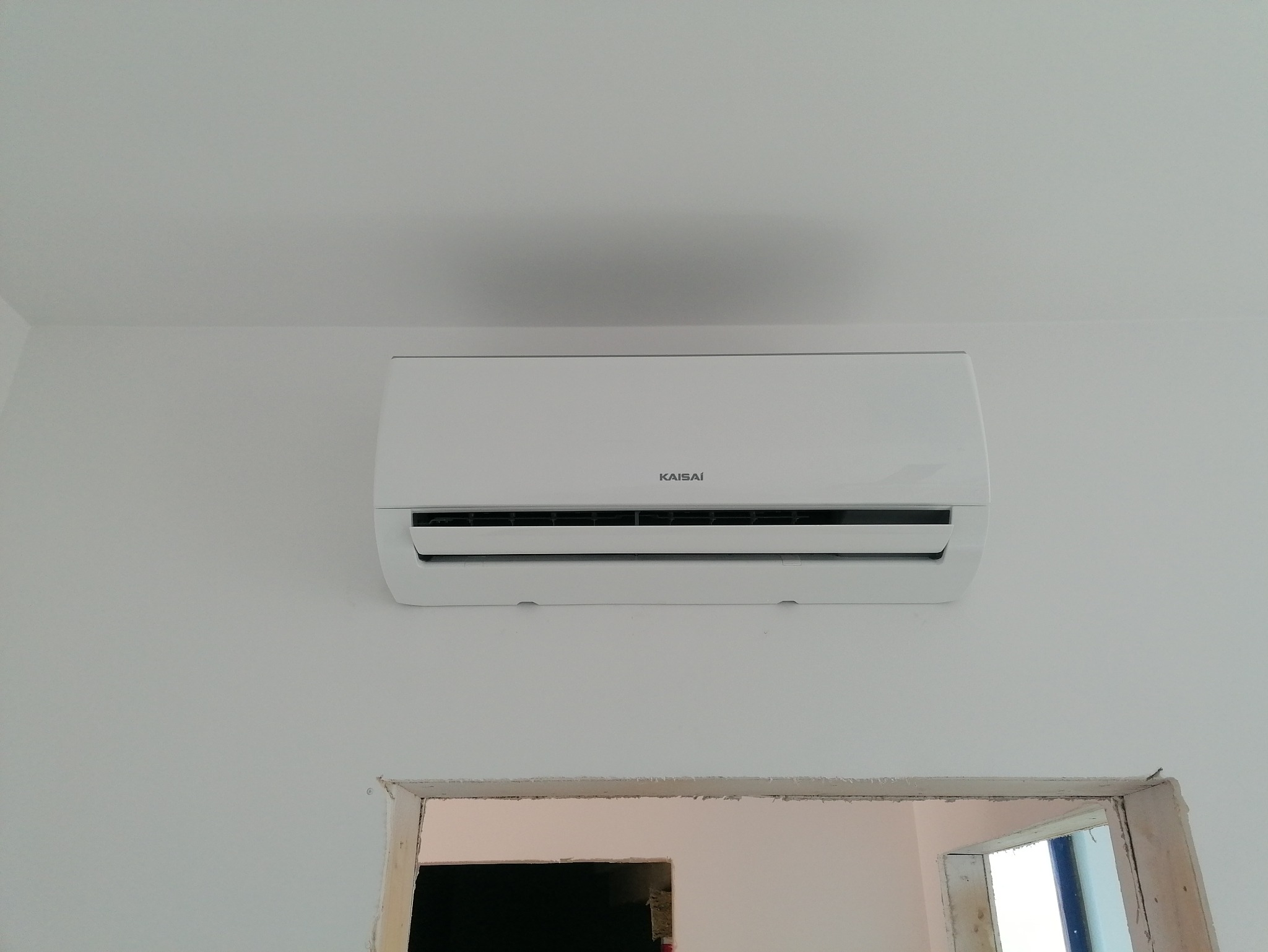 Biały klimatyzator Kaisai zamontowany na białej ścianie, widoczny fragment ościeżnicy drzwi. Minimalistyczny design, świeżo po instalacji.