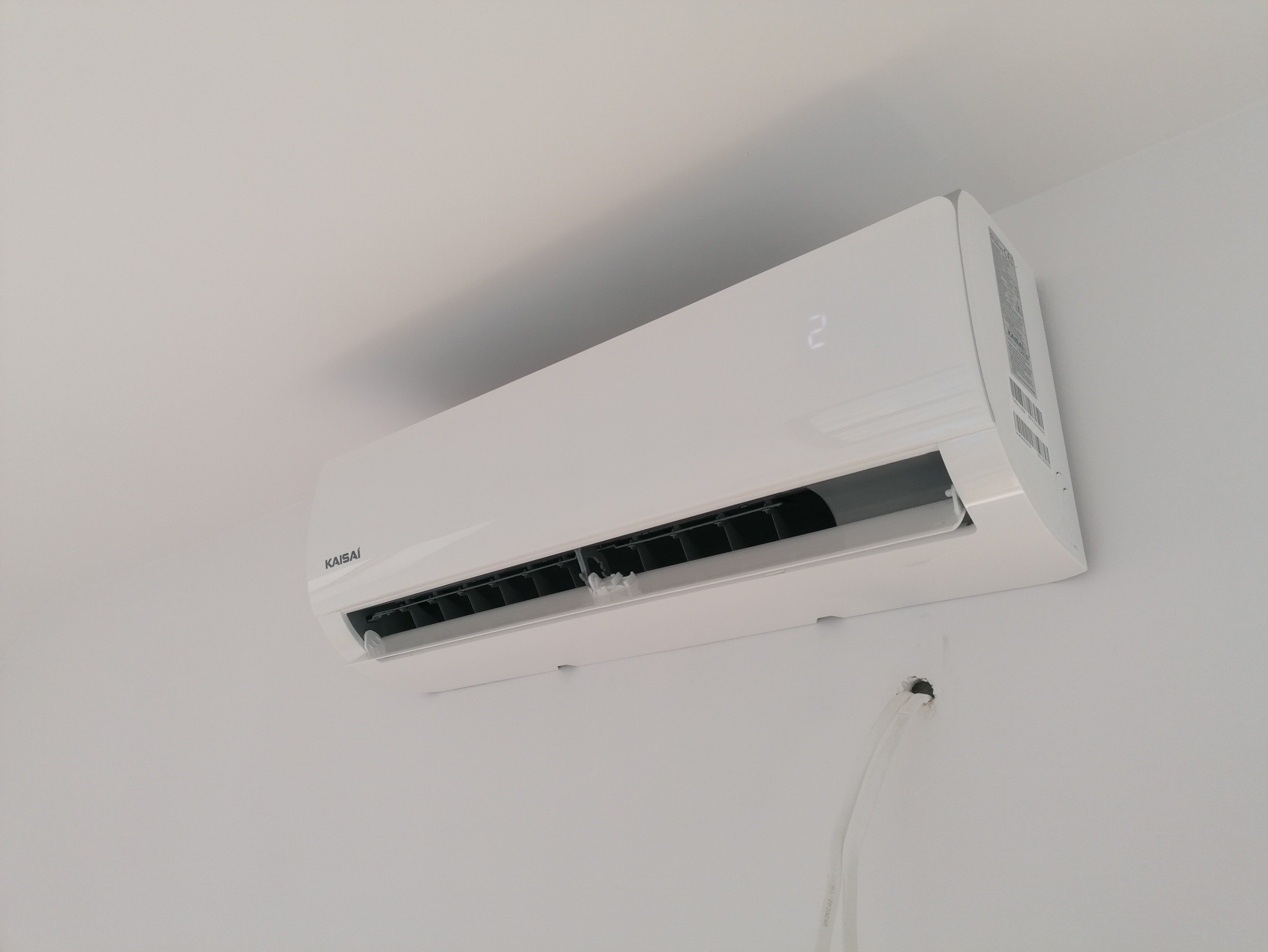 Biały klimatyzator Kaisai zamontowany na białej ścianie, widoczne rurki instalacyjne. Minimalistyczny design, nowoczesne rozwiązanie chłodzące.