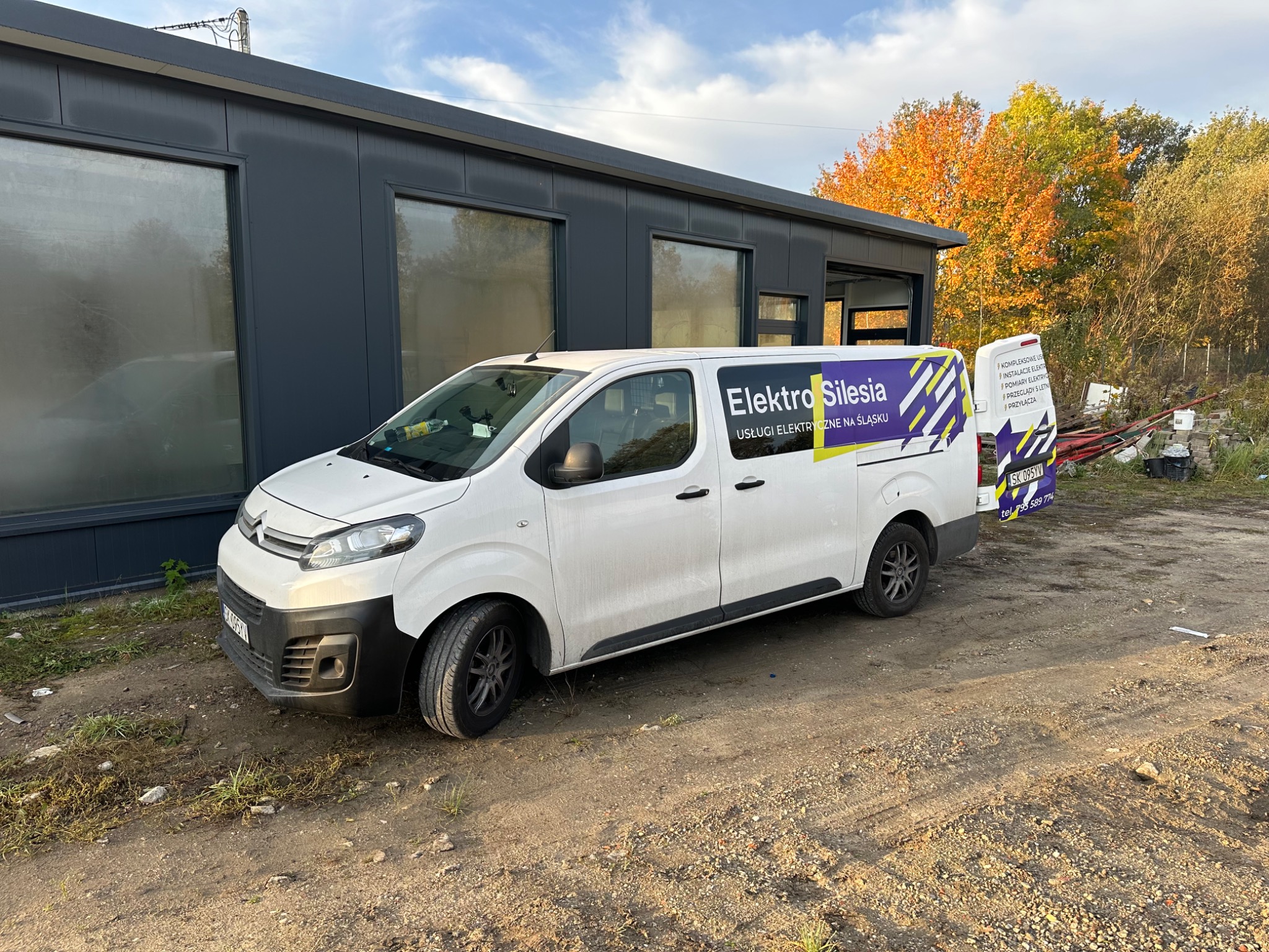 Biały samochód dostawczy z logo 'Elektro Silesia' na tle szarego budynku i drzew z jesiennymi liśćmi.