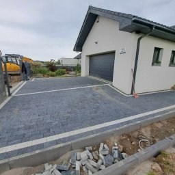SM BRUK Mateusz Suwalski - Nowa, szara kostka brukowa ułożona na podjeździe przed nowoczesnym domem z szarą bramą garażową i numerem 21, widoczny fragment wykopu i maszyna budowlana w tle.