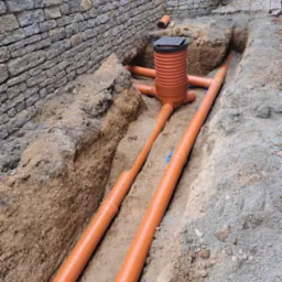 Pomarańczowe rury kanalizacyjne ułożone w wykopie, widoczna studzienka rewizyjna z pokrywą, w tle kamienny murek oporowy i fragment chodnika.